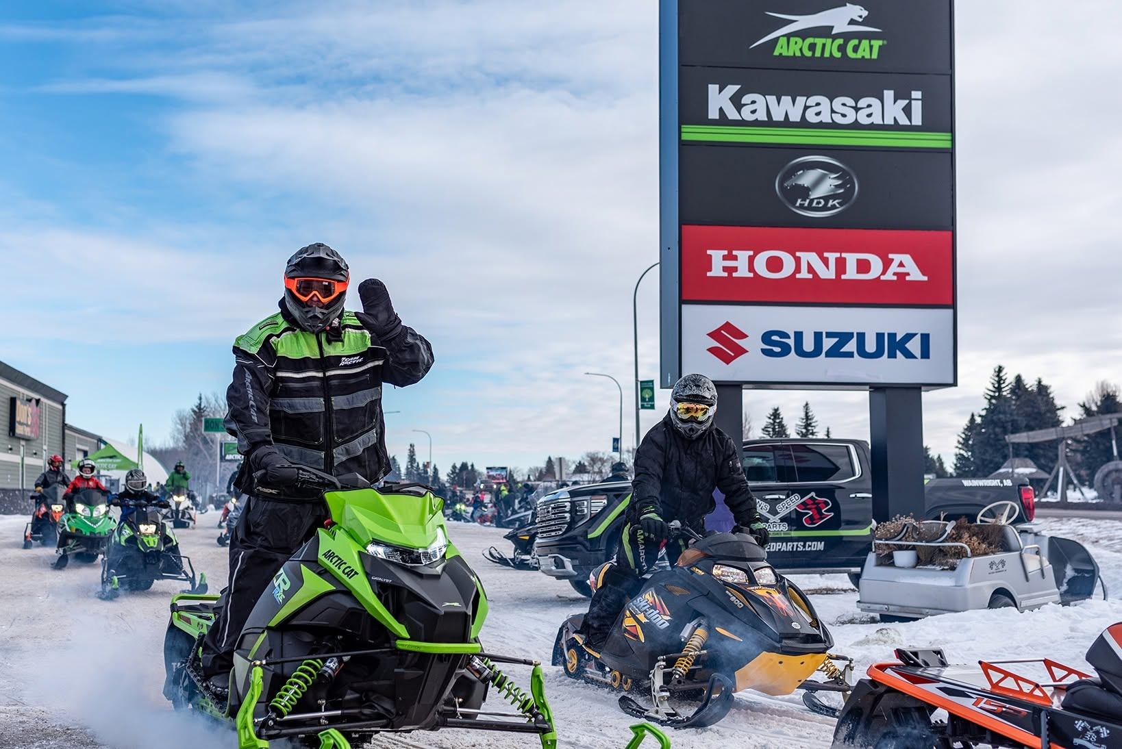 Snowmobilers gather beside Arctic Cat, Kawasaki, Honda and Suzuki dealership signs.