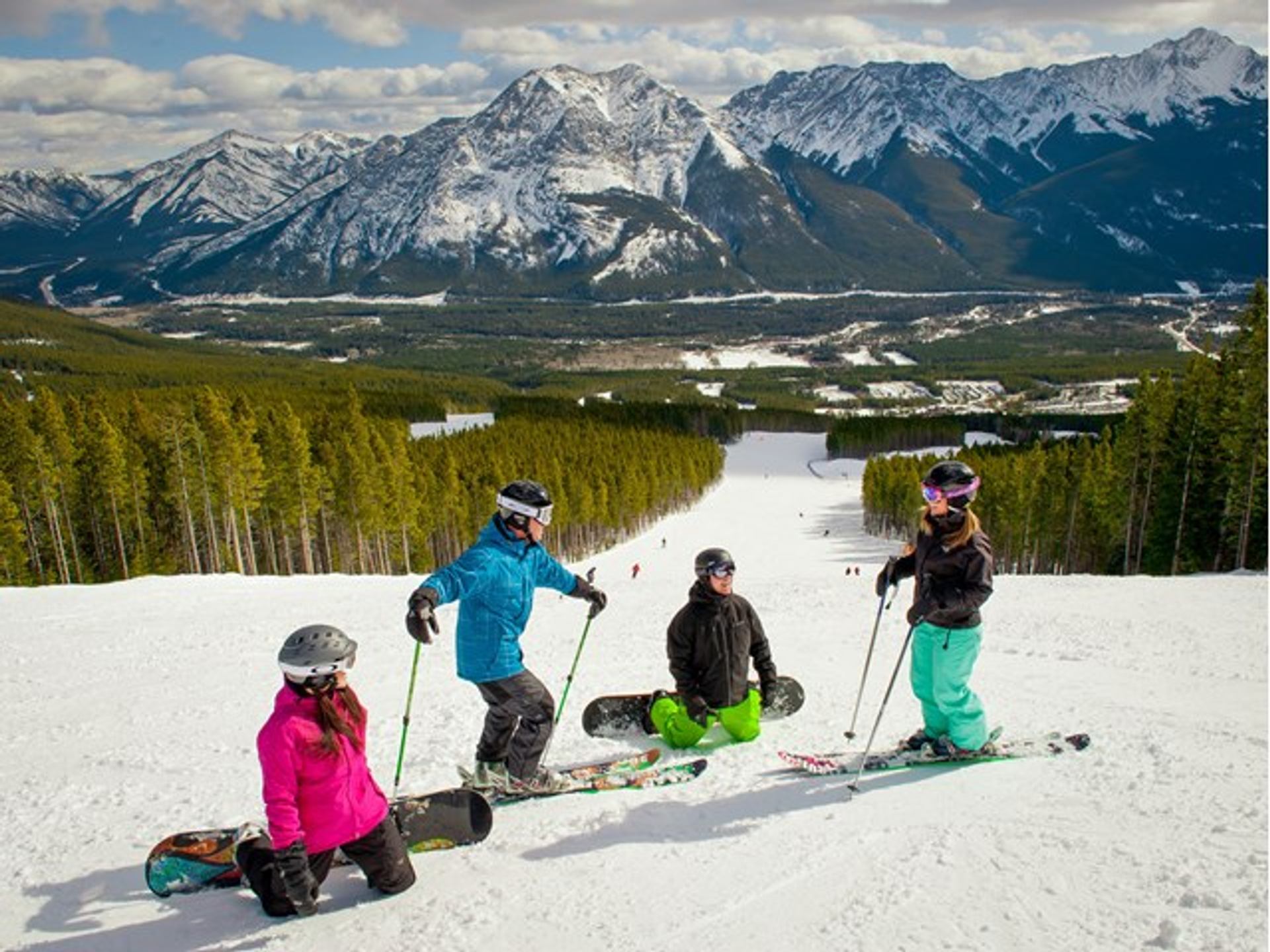 A group of four skiers and snowboarders take a break on the side of the mountain while skiing at Nakiska Ski Resort, with views of the mountains in Kananaskis Country