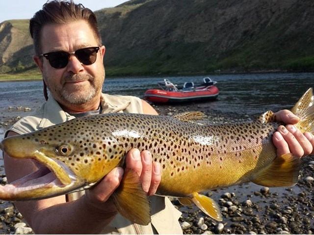 Man holds speckled trout on a rocky riverbank with red fishing boat in background.