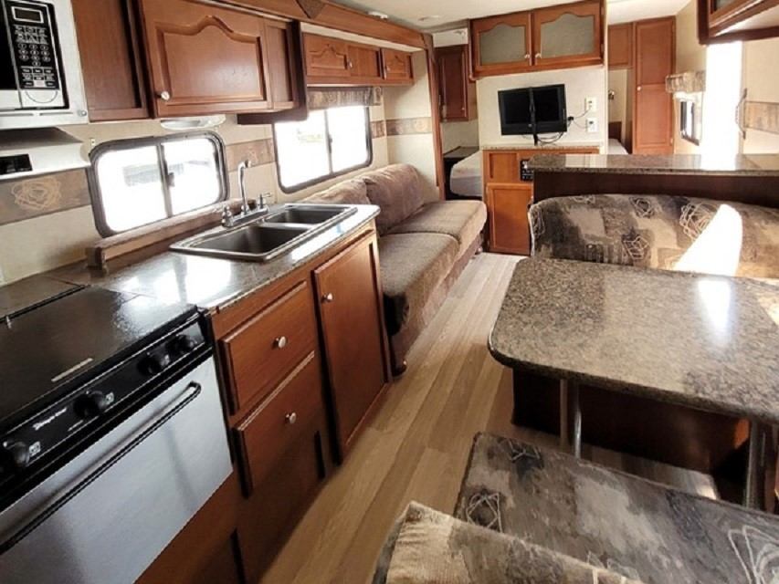 RV interior with kitchen counter, sink, stove, and bed near a flat-screen TV.
