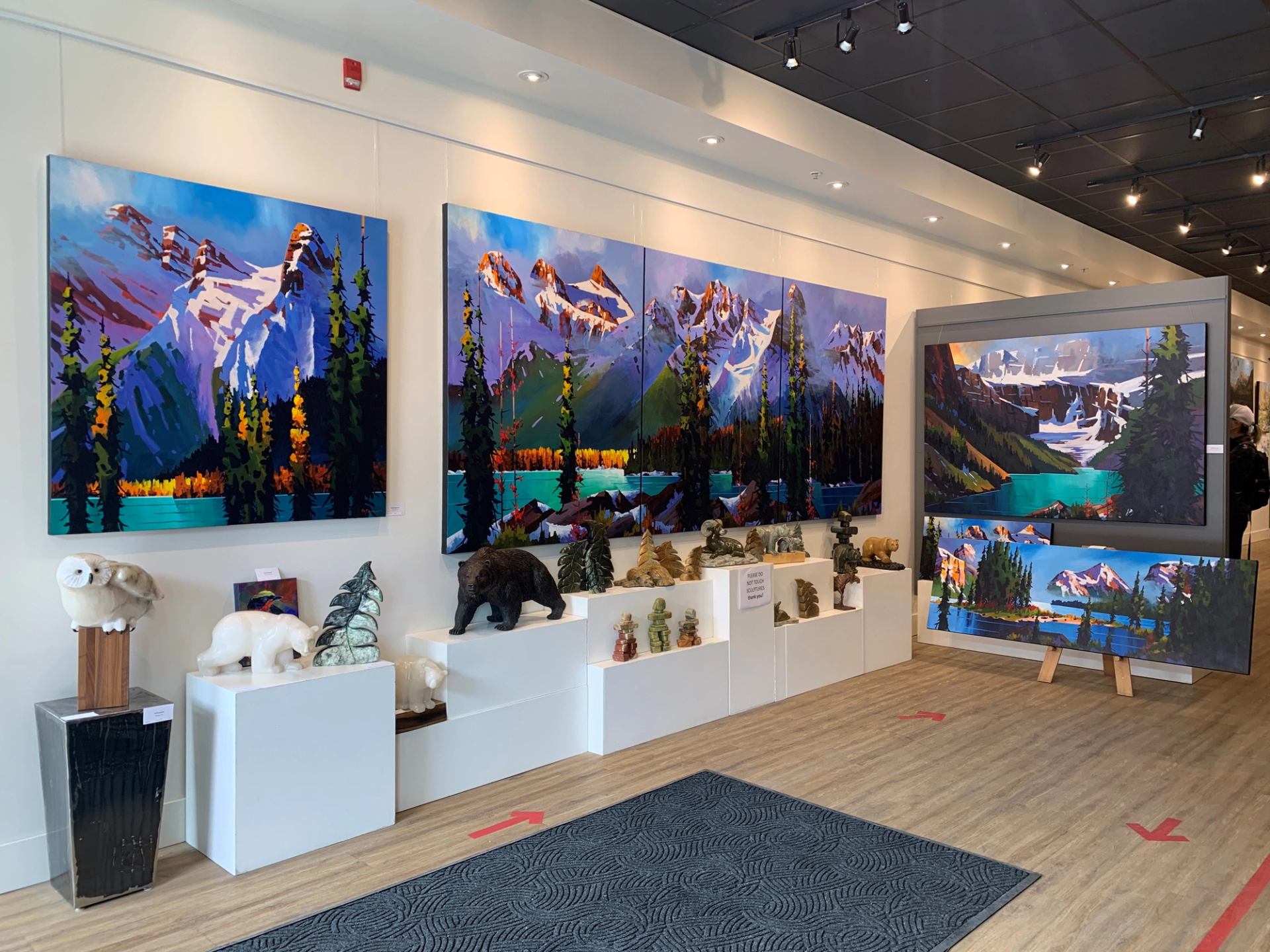 A gallery wall displaying vibrant mountain paintings with animal and nature sculptures below.