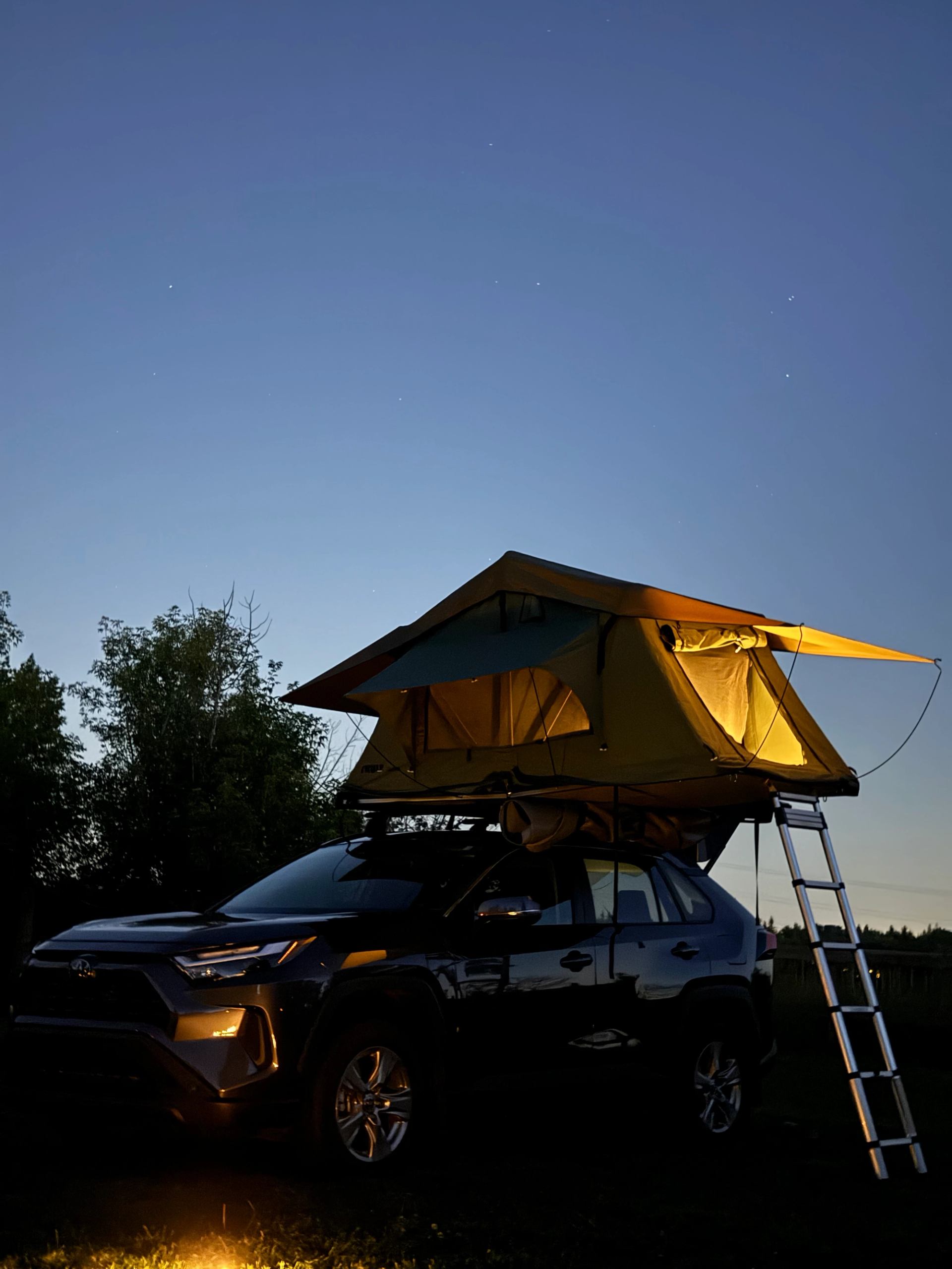 SUV with rooftop tent and ladder parked outdoors under clear evening sky.