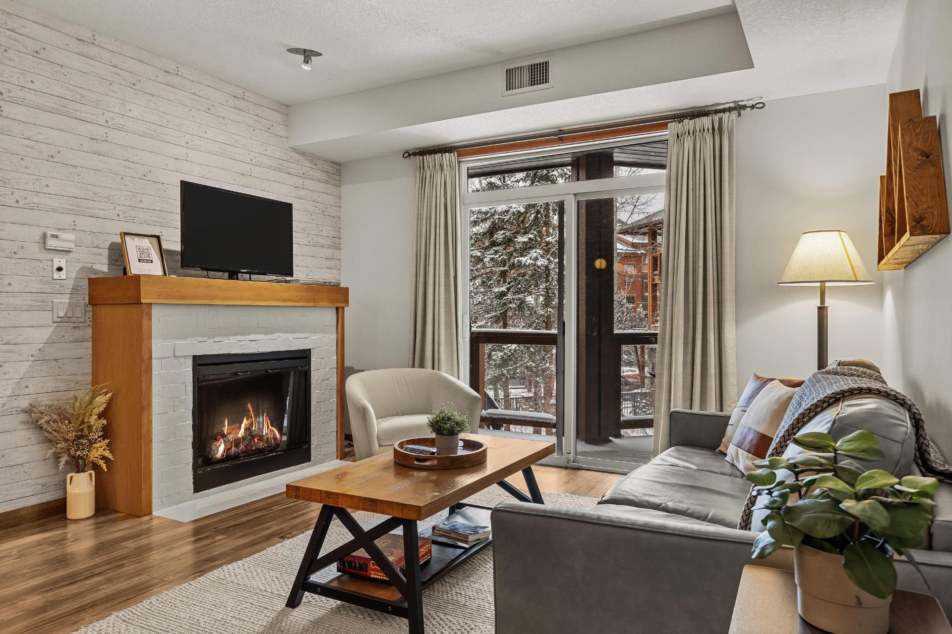 Living room with fireplace, TV, sofa, armchair and balcony overlooking snowy trees.