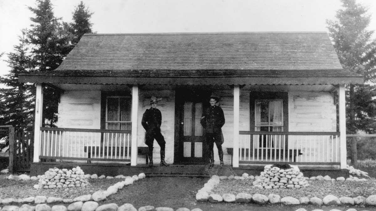 Two men in uniform stand on the porch of a small log cabin with a gabled roof and rock-lined garden paths.