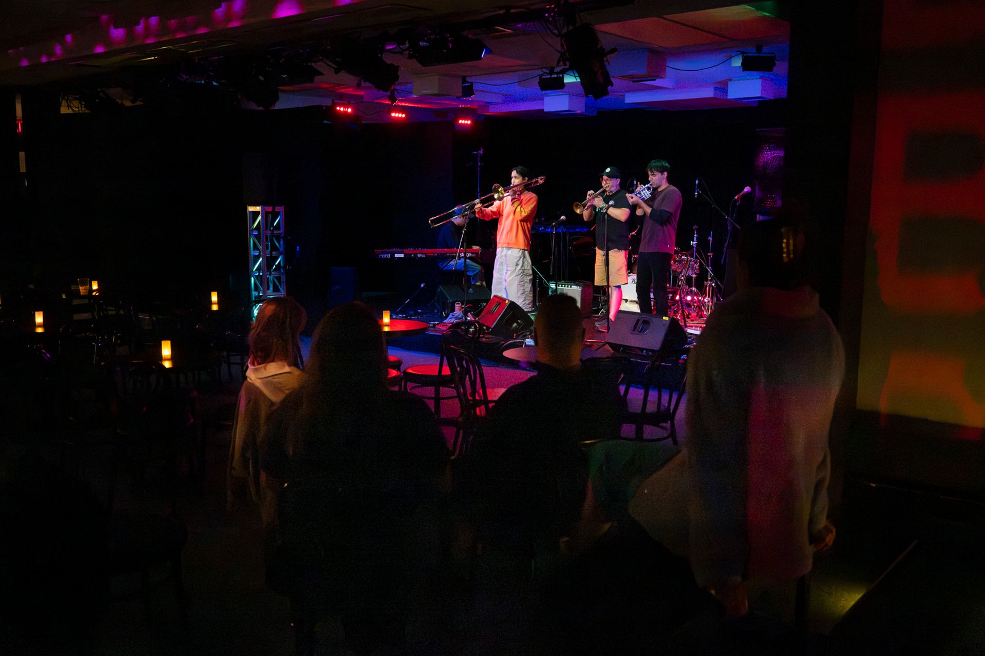 Small band plays brass instruments on stage in dimly lit club with seated audience.