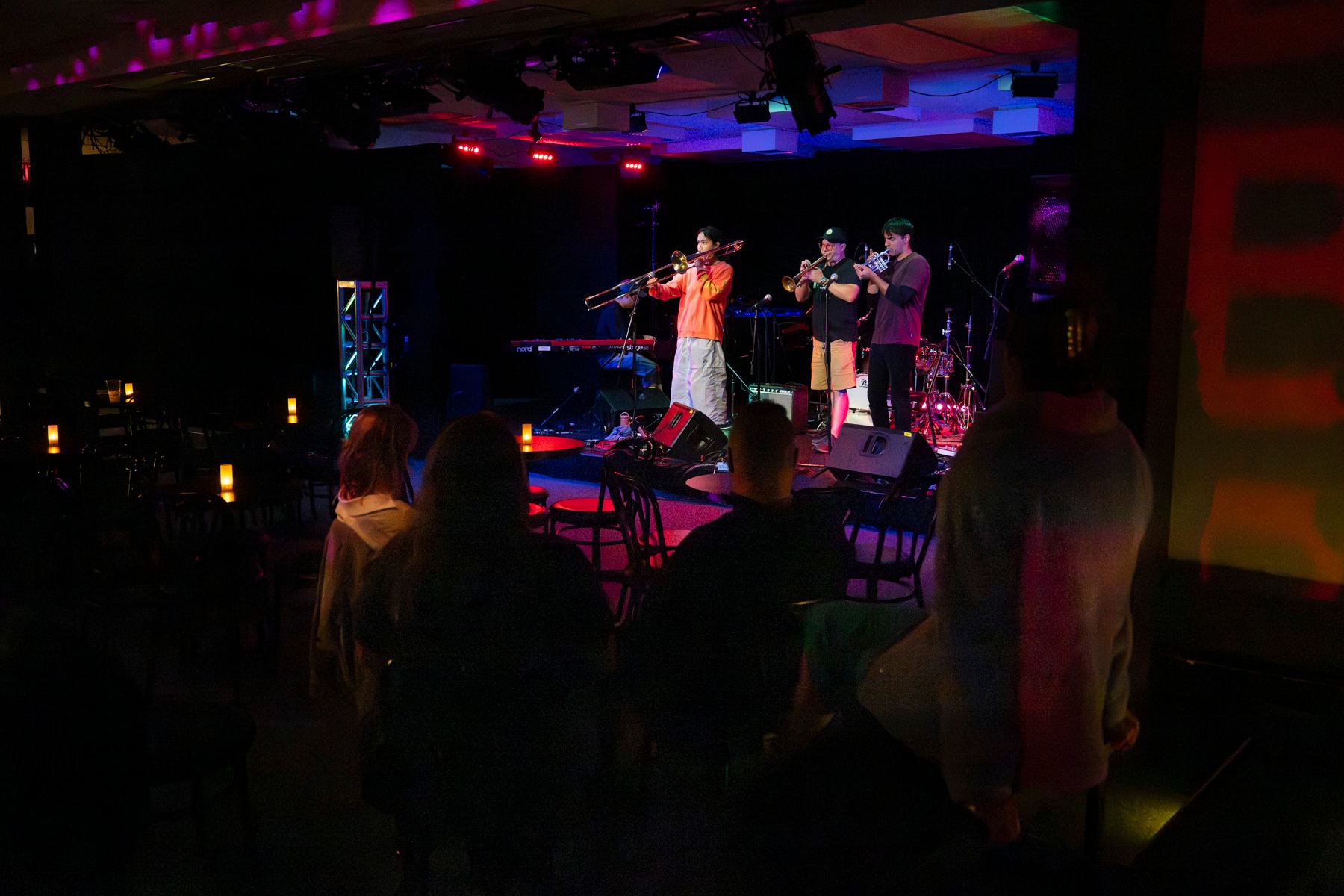Small band plays brass instruments on stage in dimly lit club with seated audience.