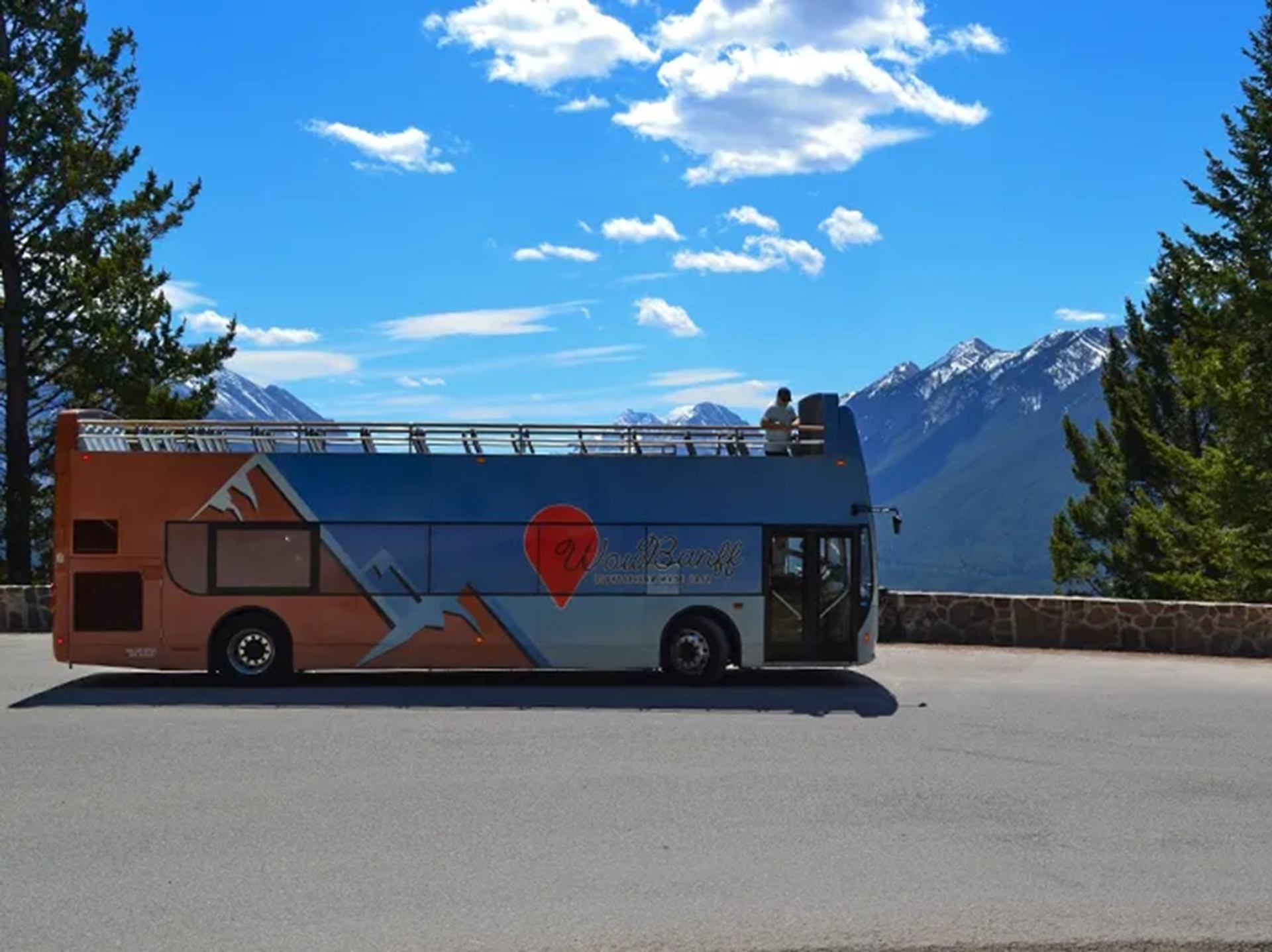Open-top tour bus parked at a mountain viewpoint beneath a blue, cloud-dotted sky.