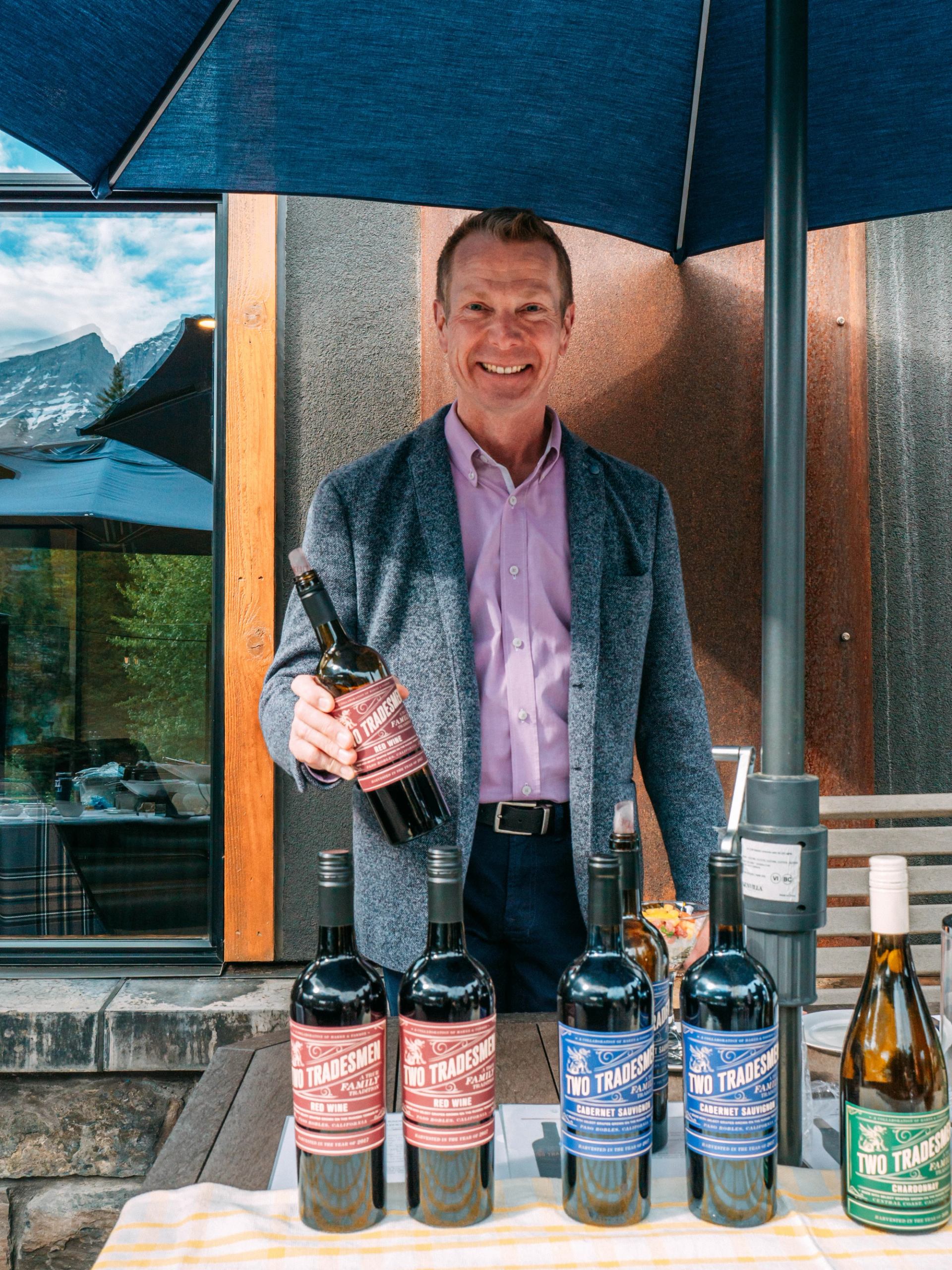 Person standing under patio umbrella holding wine bottle behind table with Two Tradesmen wines.