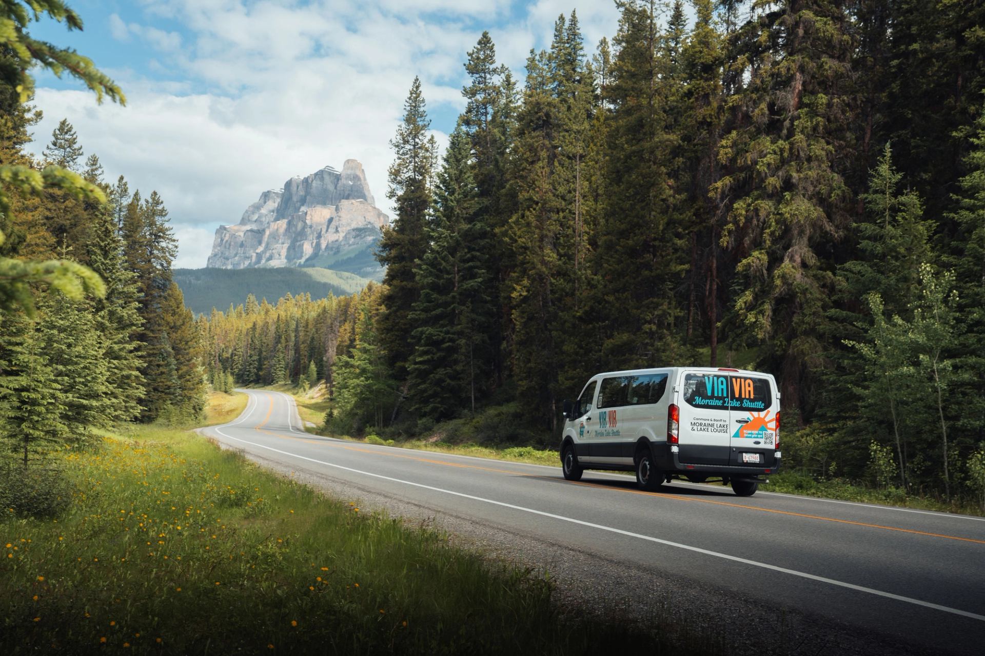 White tour van on forest road with mountain backdrop and partly cloudy sky.
