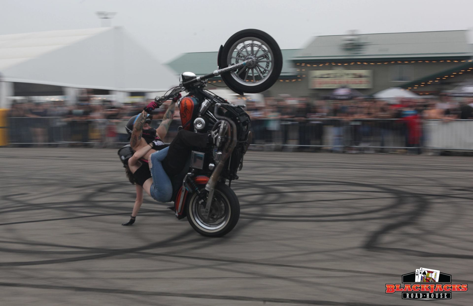 A motorcyclist performs a high-speed wheelie stunt with a passenger hanging off the back of the bike at Blackjacks Roadhouse.
