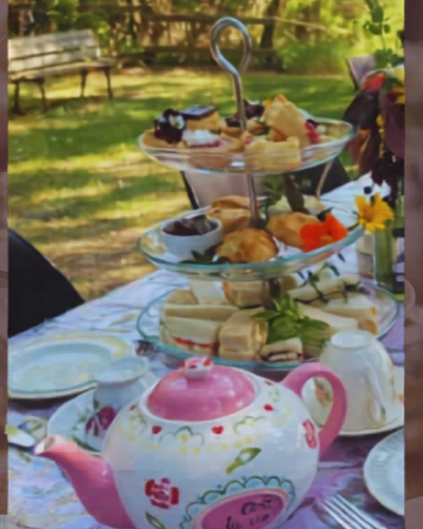 Tiered tray of pastries and sandwiches with floral teapot on outdoor garden table.