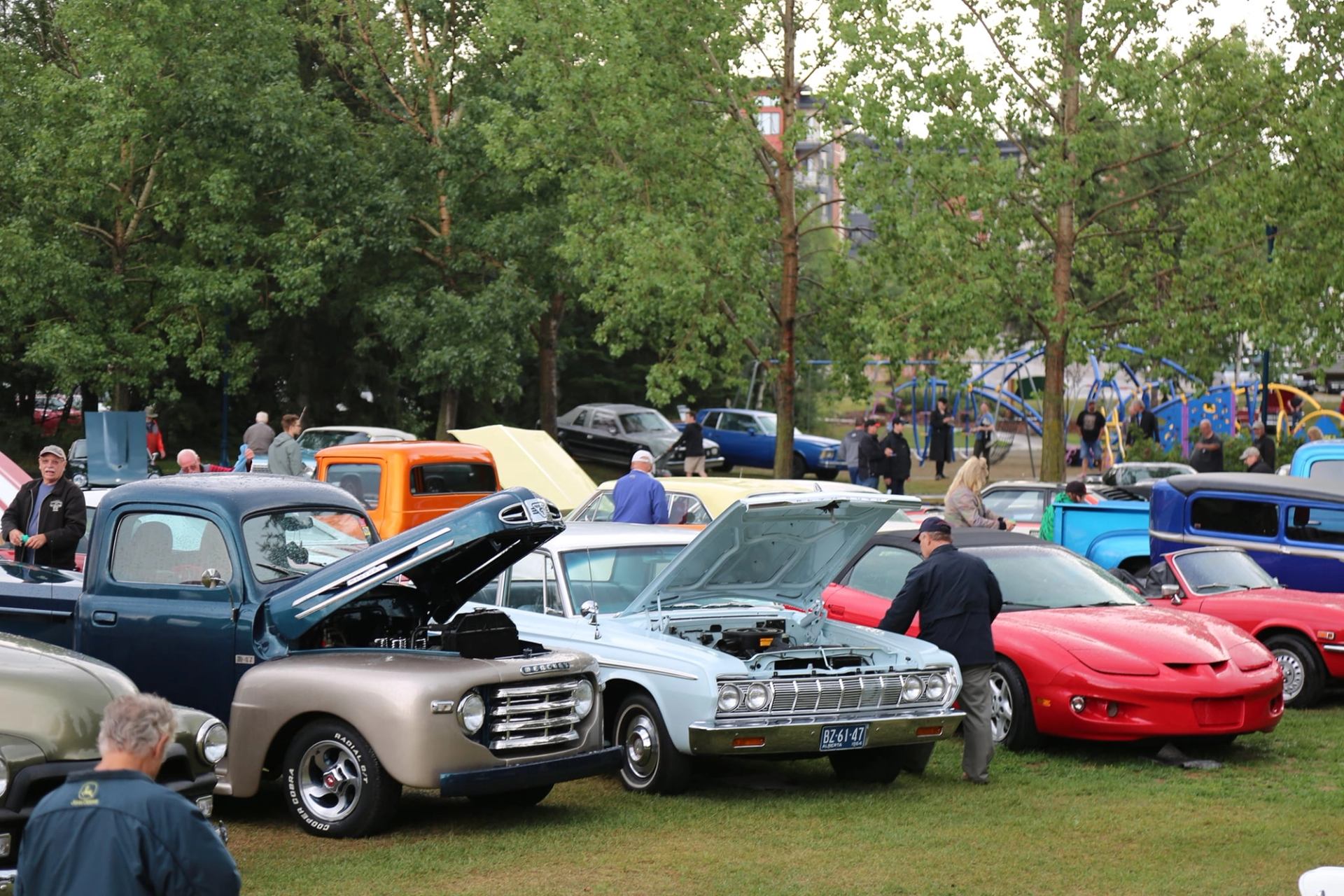 A park full of classic cars with open hoods and people attending a car show, with trees and a playground in the background.