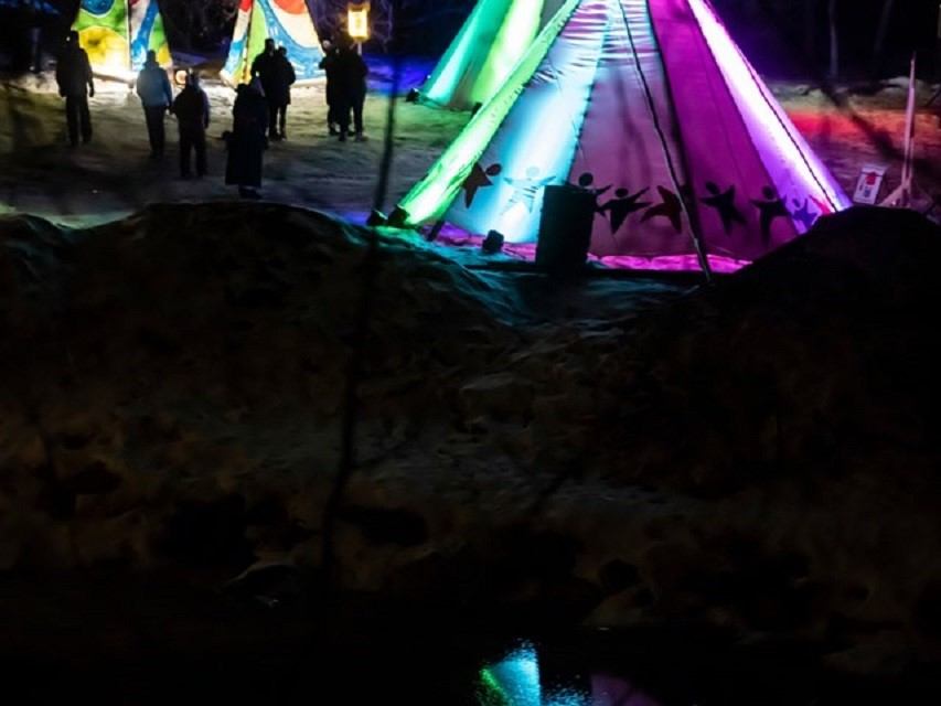 Colorful lit teepees glow at night with people walking around at winter festival.