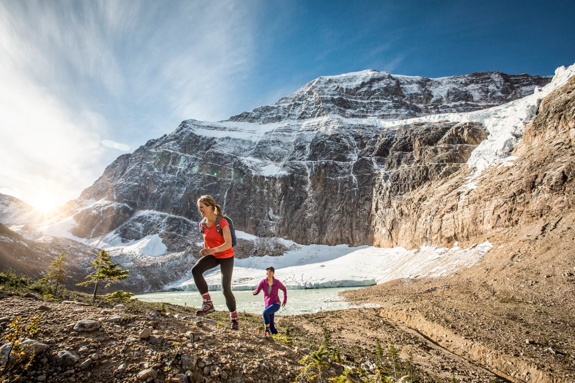 Two women hike at Mount Edith Cavell, with Cavell Pond, Cavell Glacier, and Angel Glacier behind in Jasper National Park.