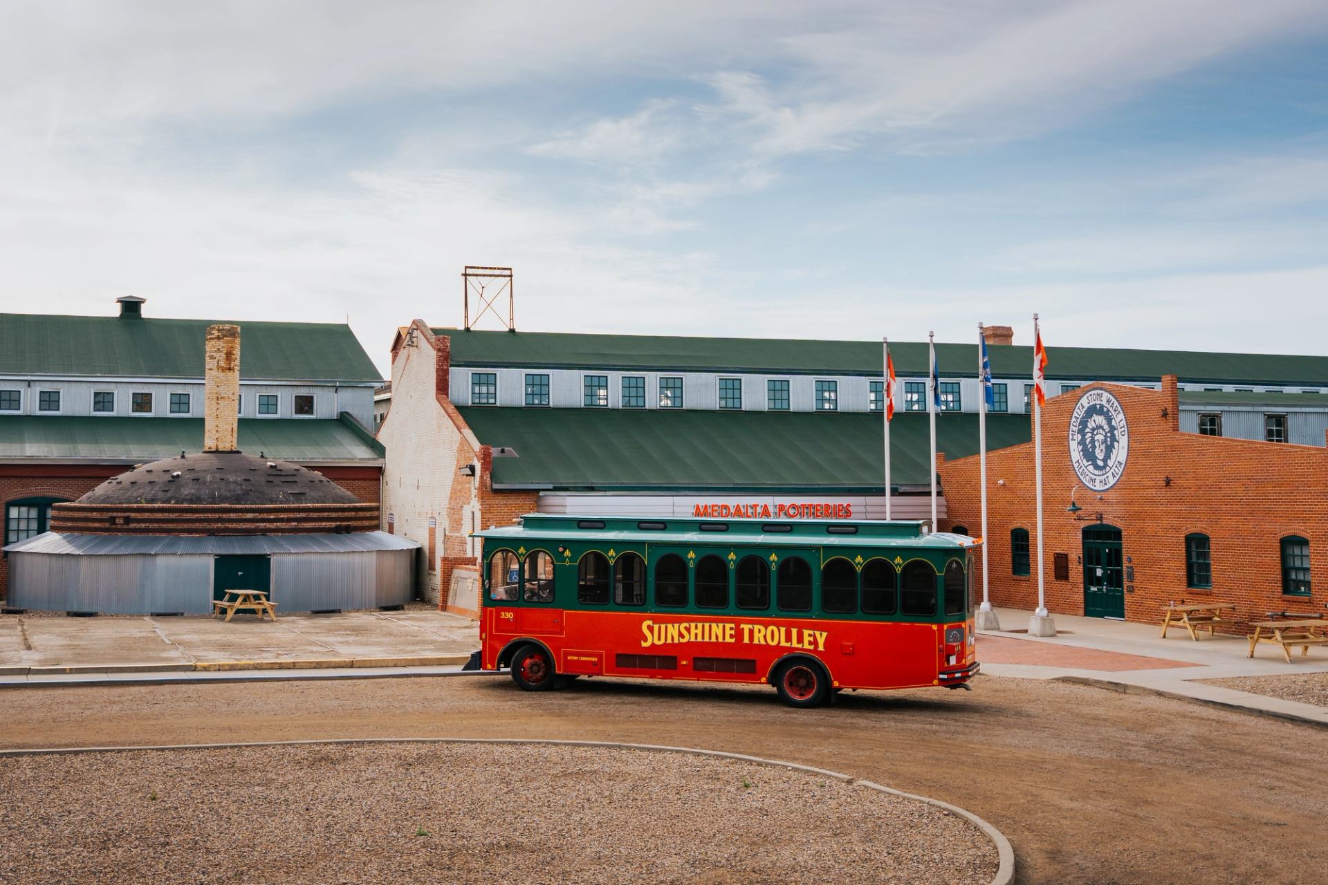 Red and green Sunshine Trolley parked outside Medalta Potteries brick industrial buildings.