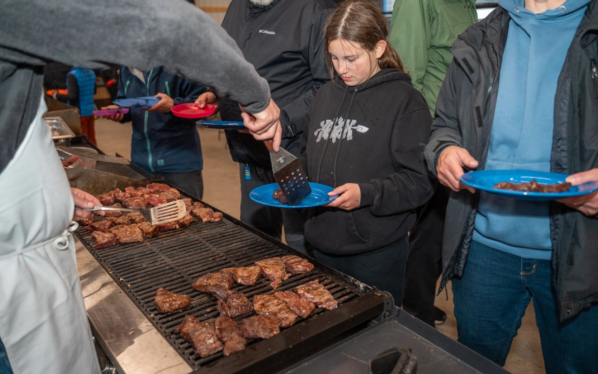 People lining up at a grill to receive cooked meat during a group meal.