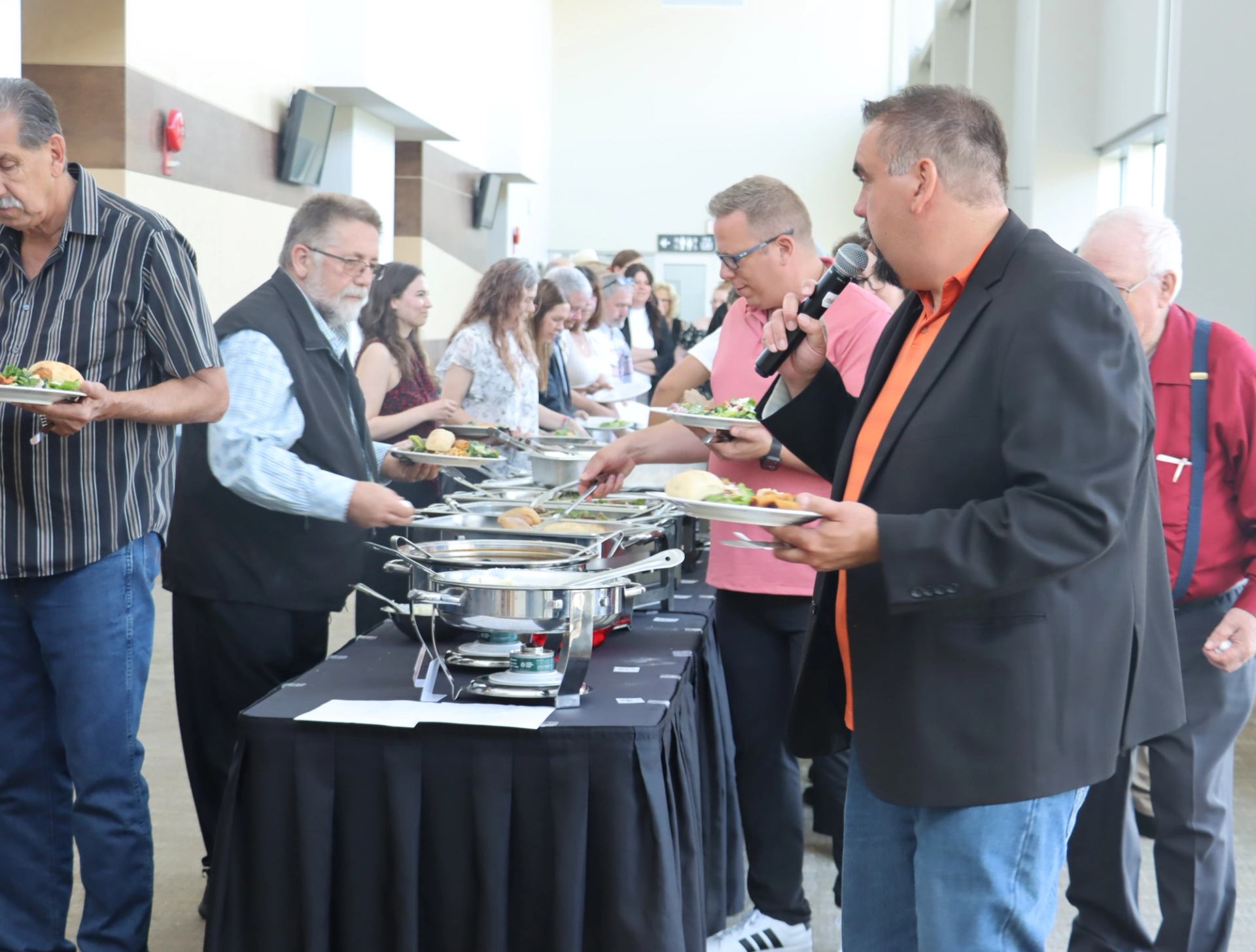People serve themselves food from a buffet table while a person speaks into microphone.