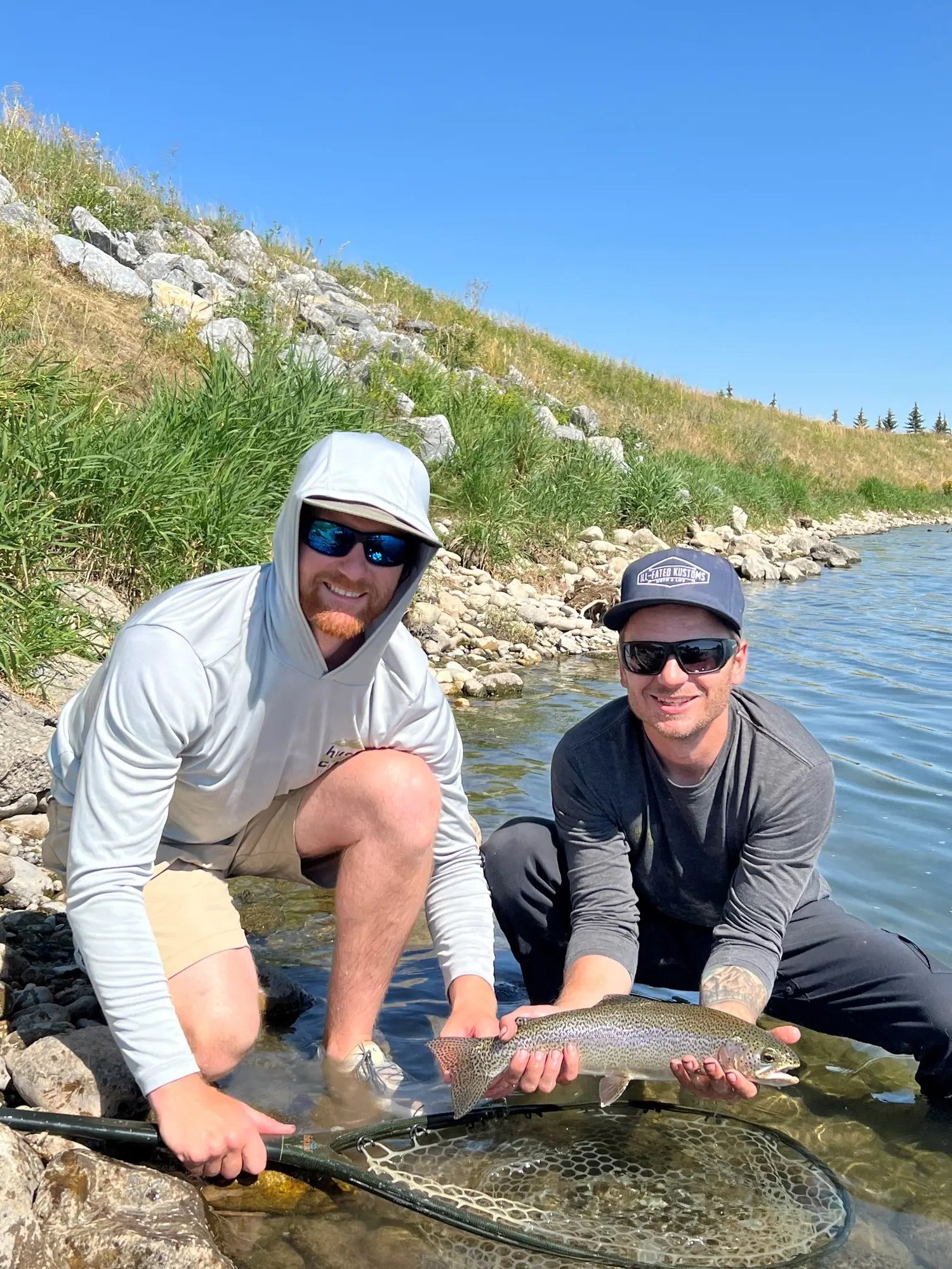 Two people kneel by a rocky shoreline, holding a trout above a landing net.