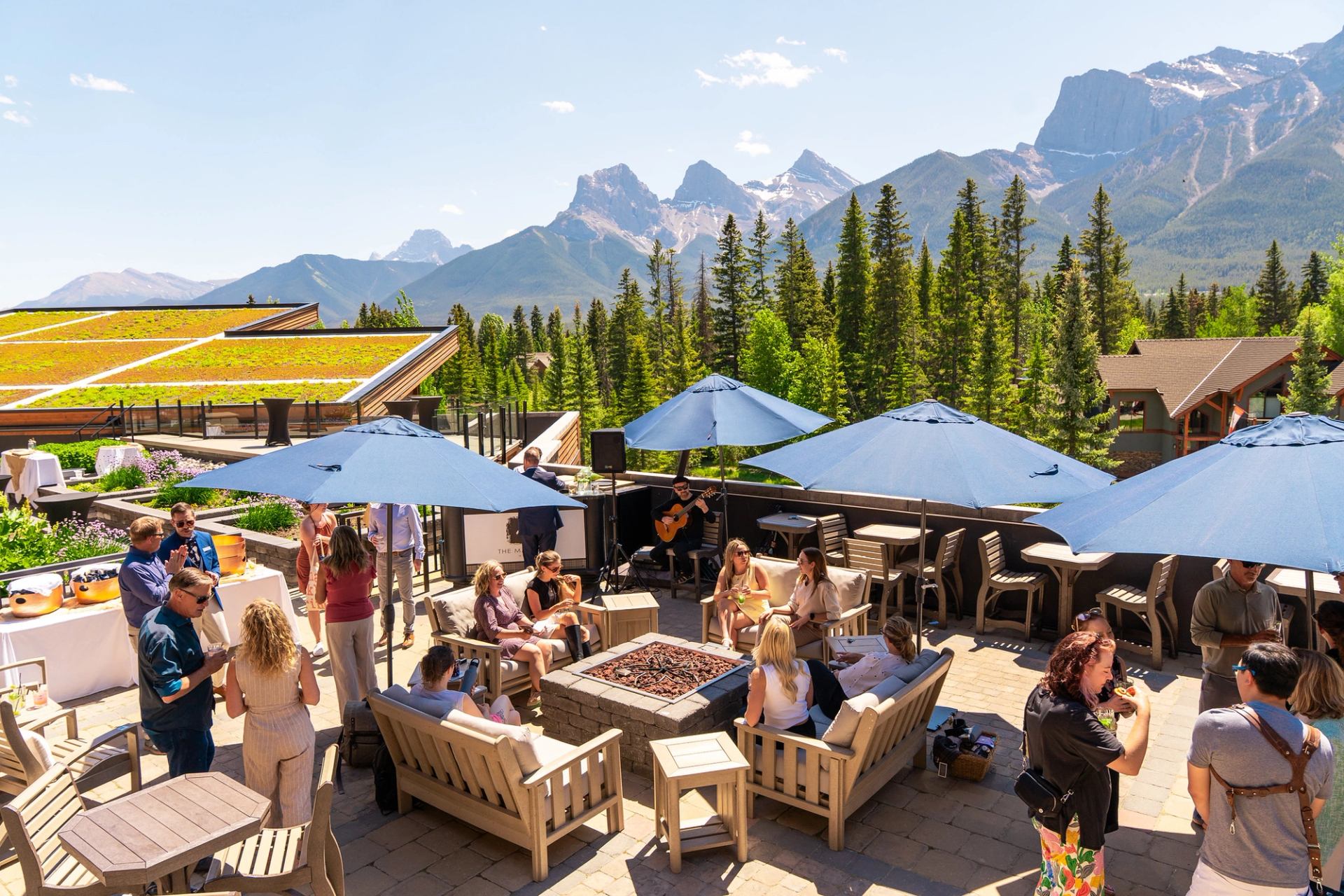 Rooftop patio gathering with blue umbrellas, firepit seating and mountain forest backdrop.