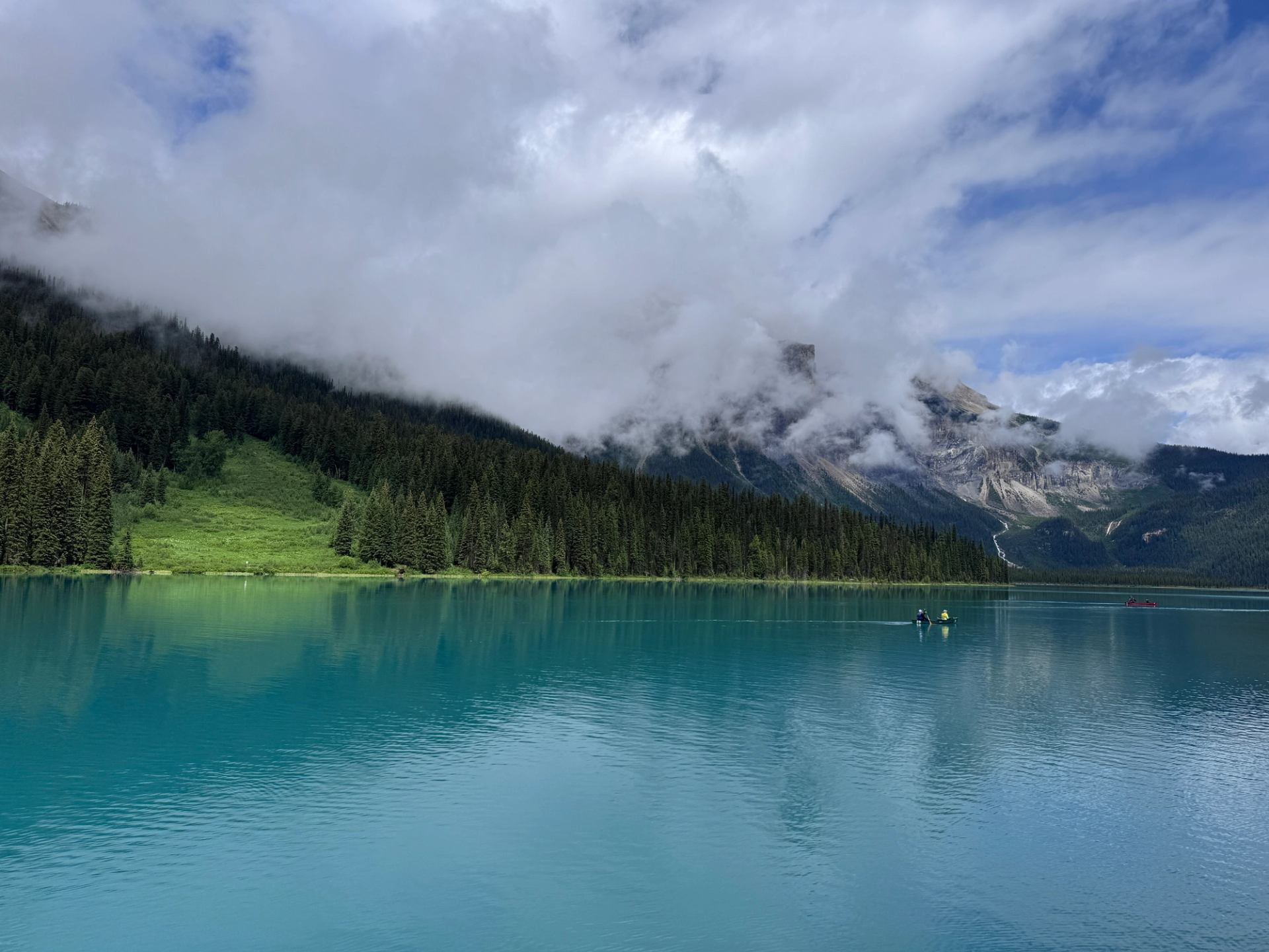 Turquoise lake with misty clouds over forested slopes and rugged mountain peaks.