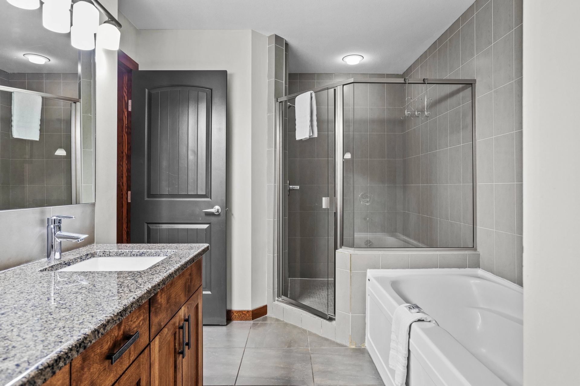 Modern bathroom with granite vanity, glass shower, bathtub and grey tiled walls.