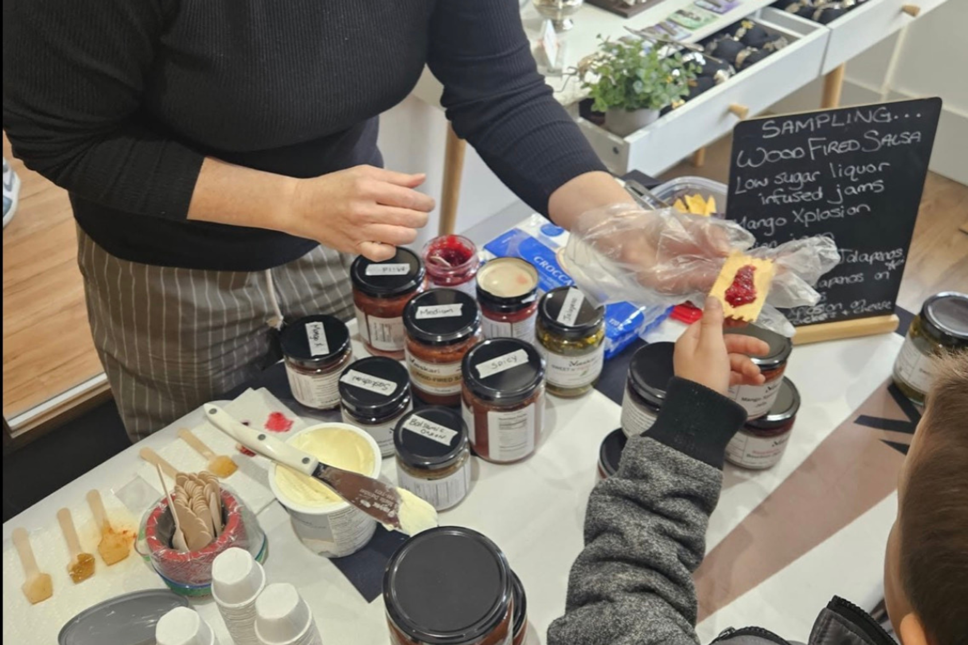 Customer samples jam on a cracker beside jars and a wood-fired salsa sign.