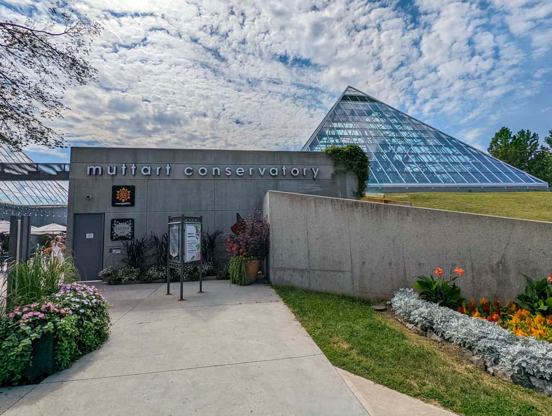 Entrance to Muttart Conservatory with glass pyramid, concrete walls and colourful flower beds.