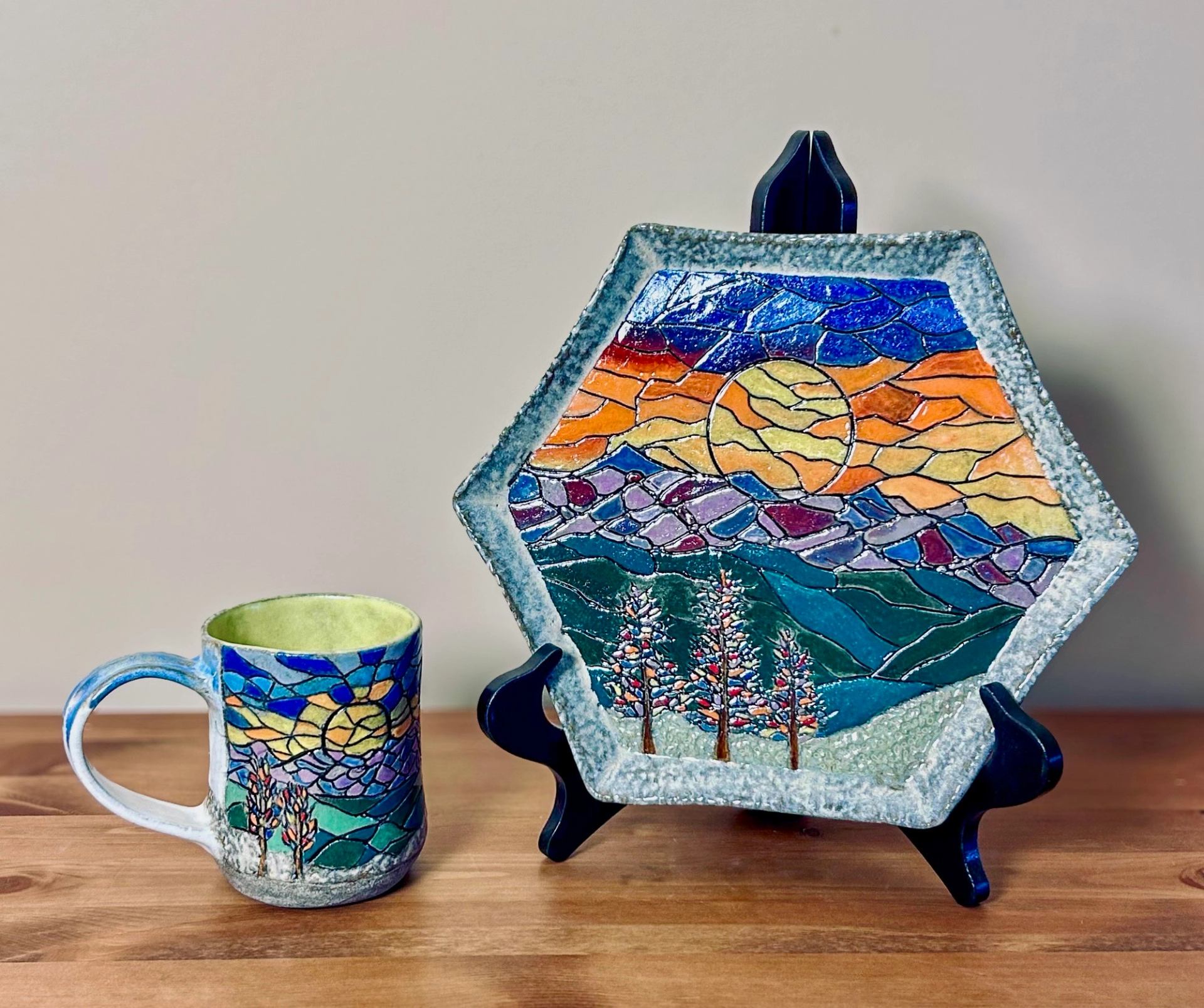 Matching ceramic mug and hexagonal plate with a vibrant stained-glass mosaic design of a sunset over mountains and trees.
