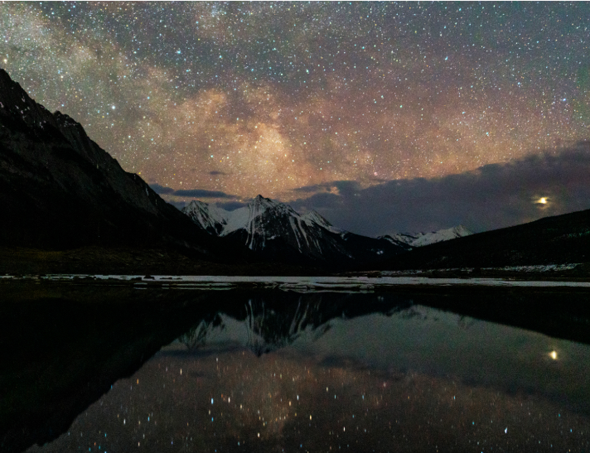 Star-filled sky over snow-capped mountains reflected in a calm, dark lake at night.