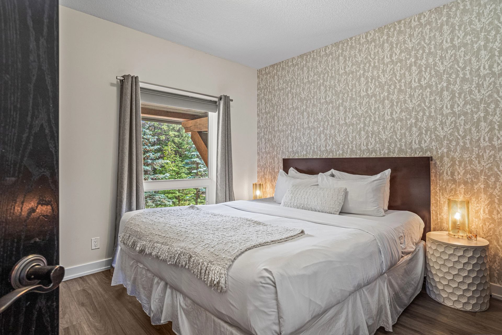Bedroom with white bedding, patterned wallpaper, bedside lamps and a window overlooking trees.