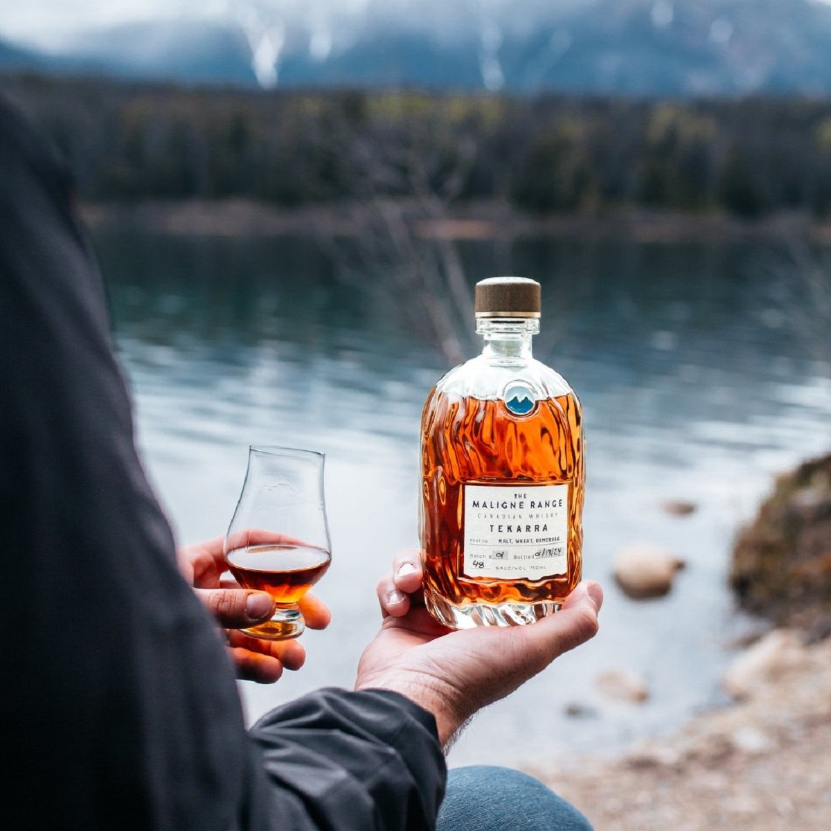 Person holding whiskey bottle and glass by a lake with mountain backdrop.