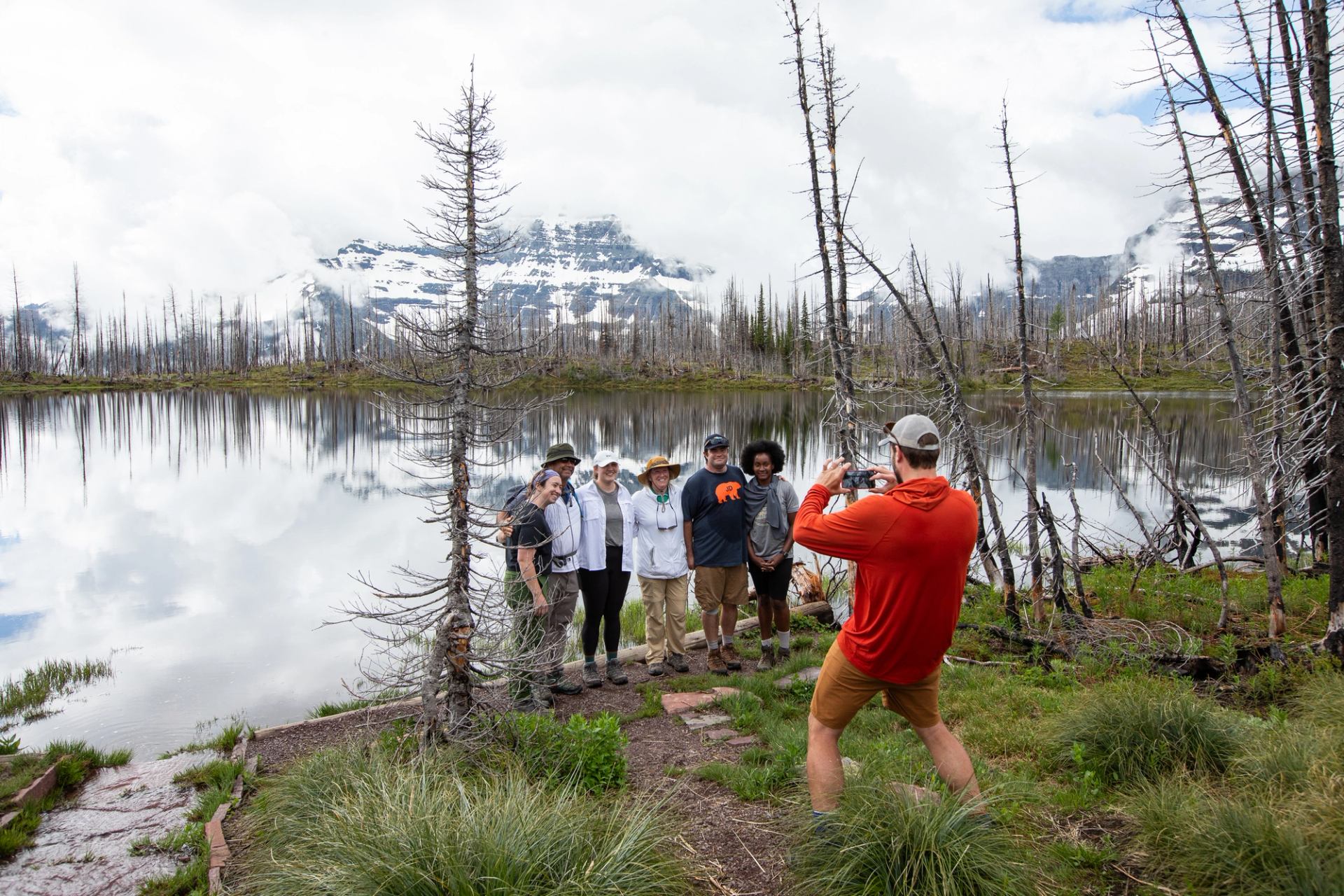 Hiker photographs group posing by reflective mountain lake surrounded by burnt trees and grass.