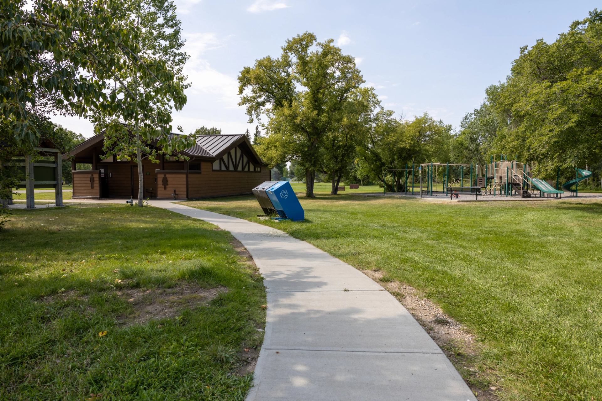 Park scene with playground, trees, recycling bins and path leading to brown building.