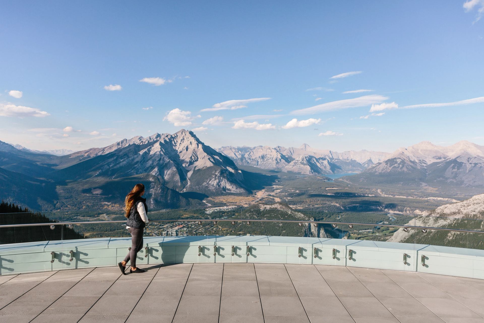 Person standing on mountain viewing platform overlooking distant peaks, valleys and blue sky.