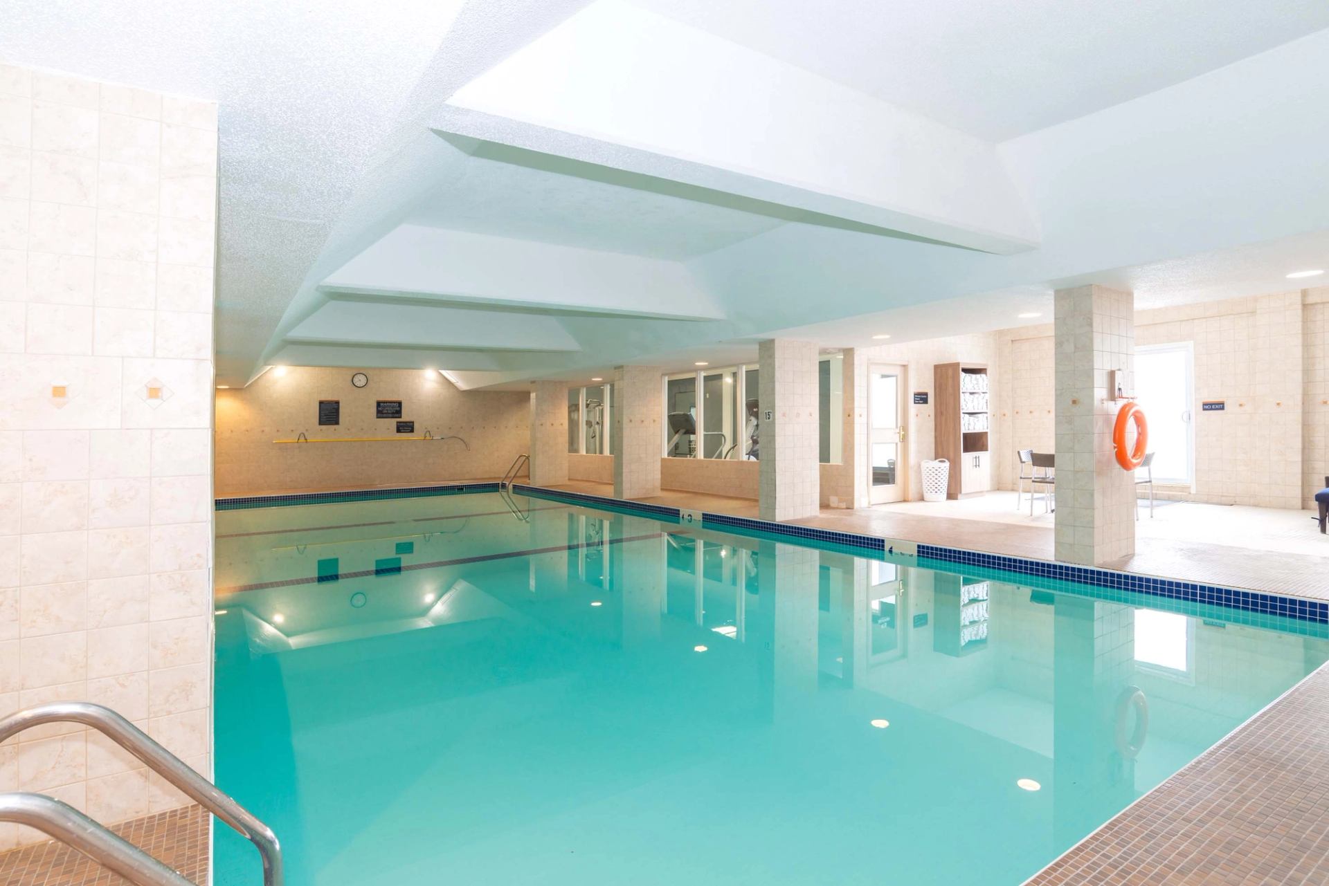 Indoor swimming pool area with seating, handrails, and bright overhead lighting.