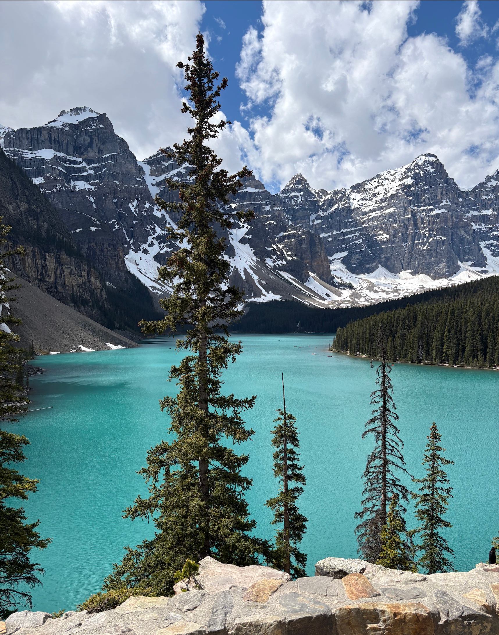 Turquoise mountain lake with evergreen trees, rocky shoreline and snow-capped peaks under clouds.