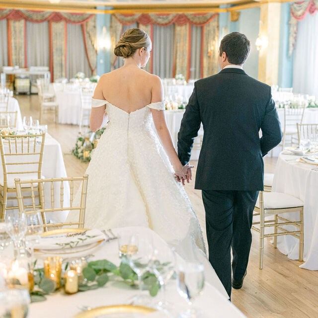 Wedding couple holding hands while walking into an elegant indoor reception with decorated tables and chairs.
