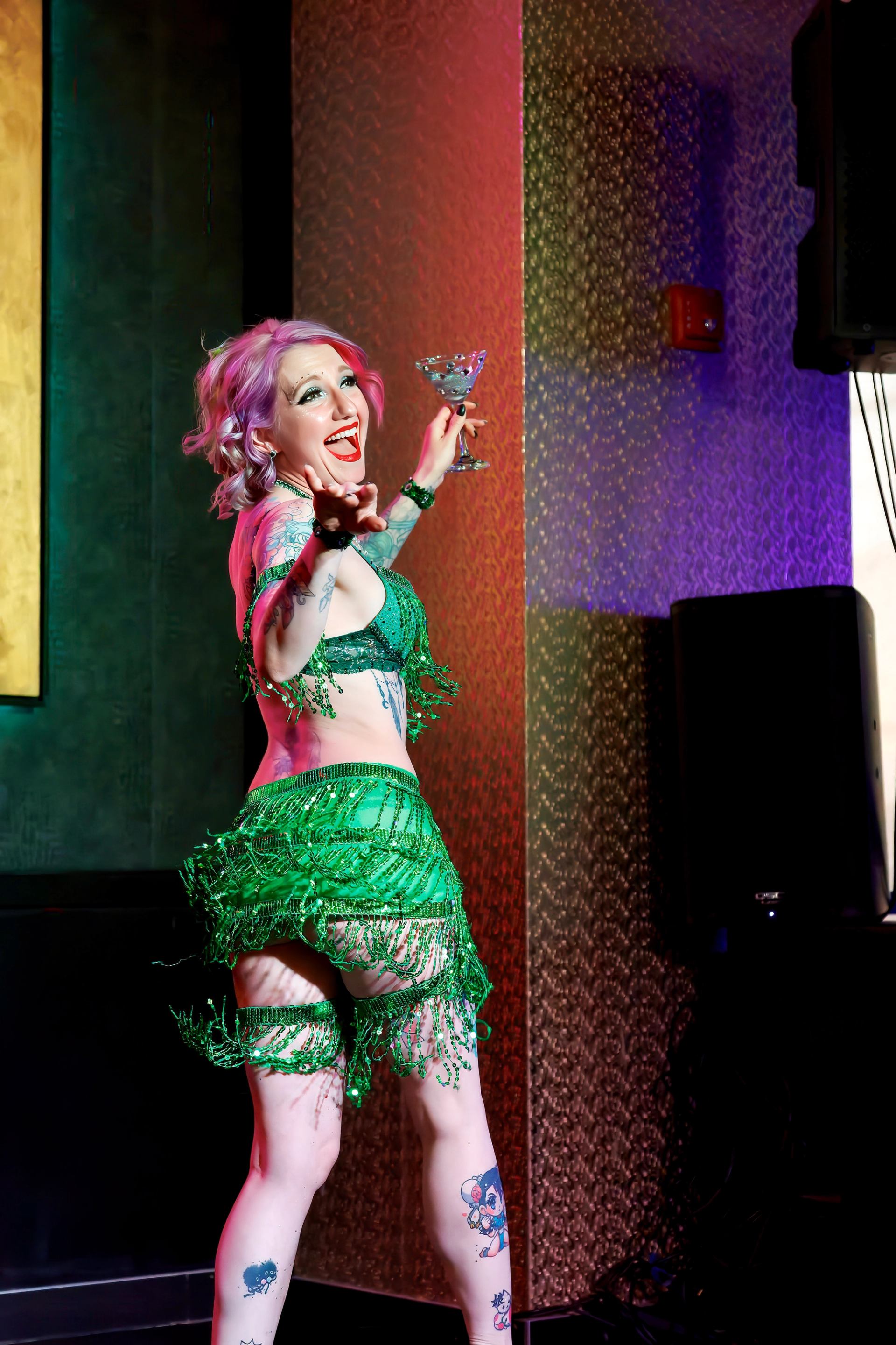 Performer in green beaded costume holds cocktail glass on stage with colourful lighting.