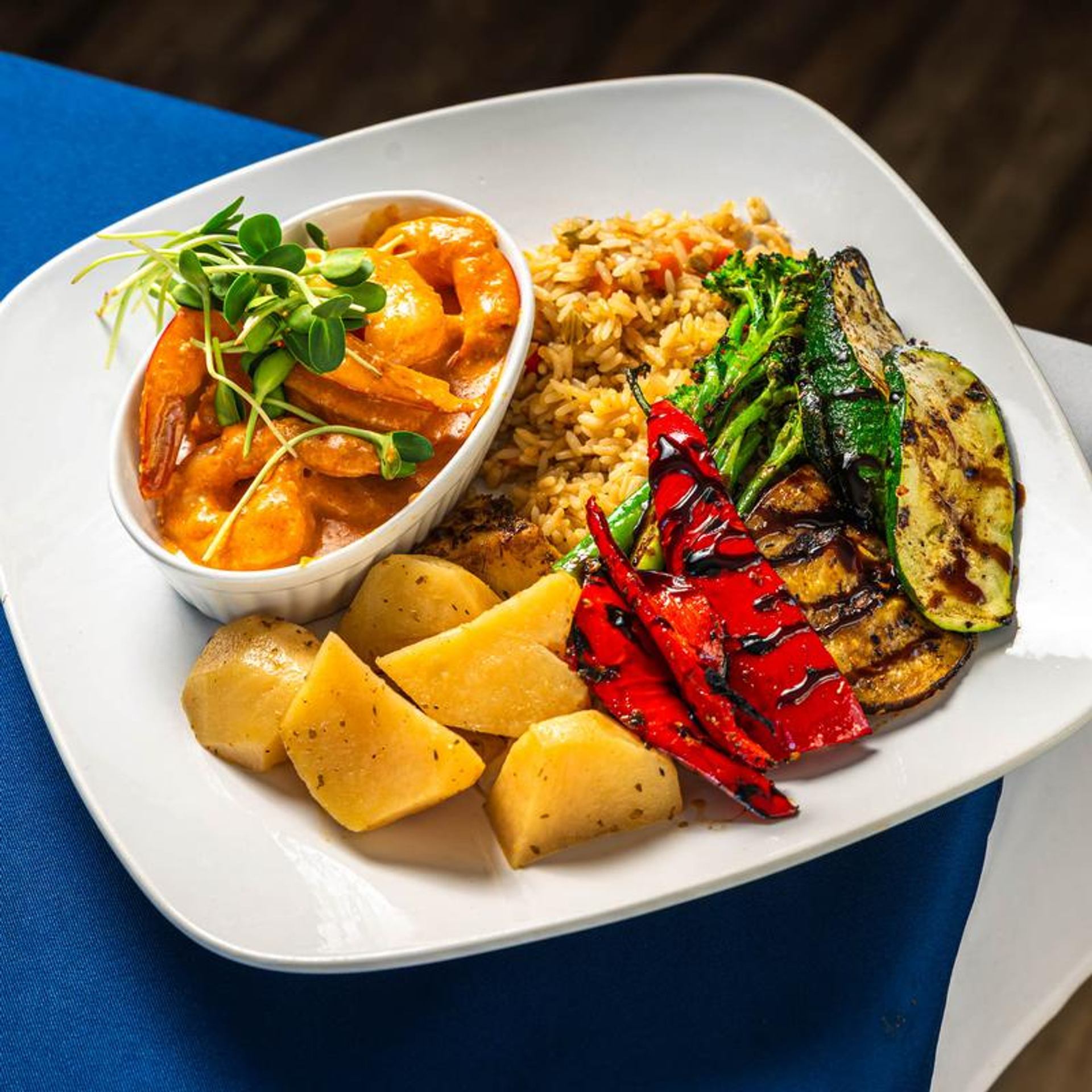 A Greek seafood dish is served with rice, grilled vegetables, potatoes, and herbs on a white plate.