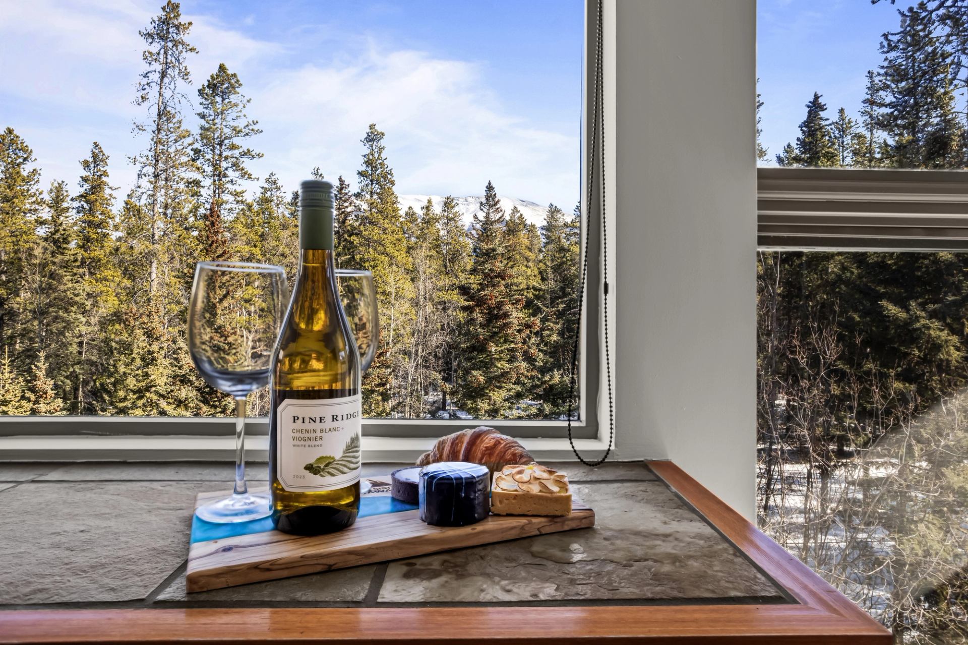 A bottle of wine, two glasses, and a small charcuterie board are arranged on a windowsill overlooking evergreen trees and snow-capped mountains in the distance.