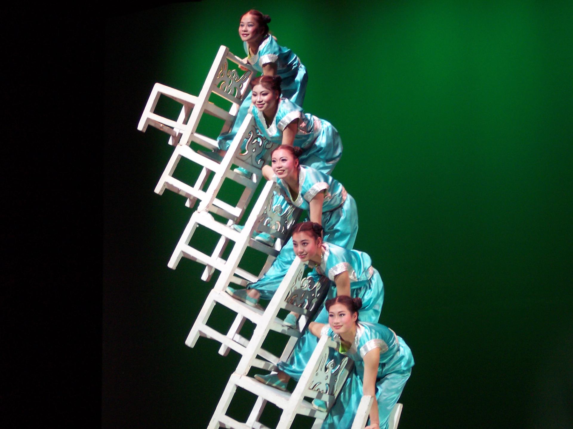 Acrobats in teal costumes balance on stacked white chairs against a green stage backdrop.