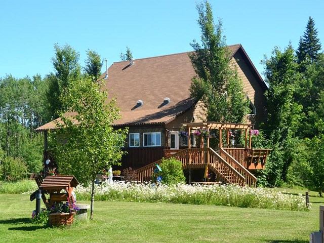 Charming countryside house with wood deck, garden, and lush greenery under a clear blue sky.