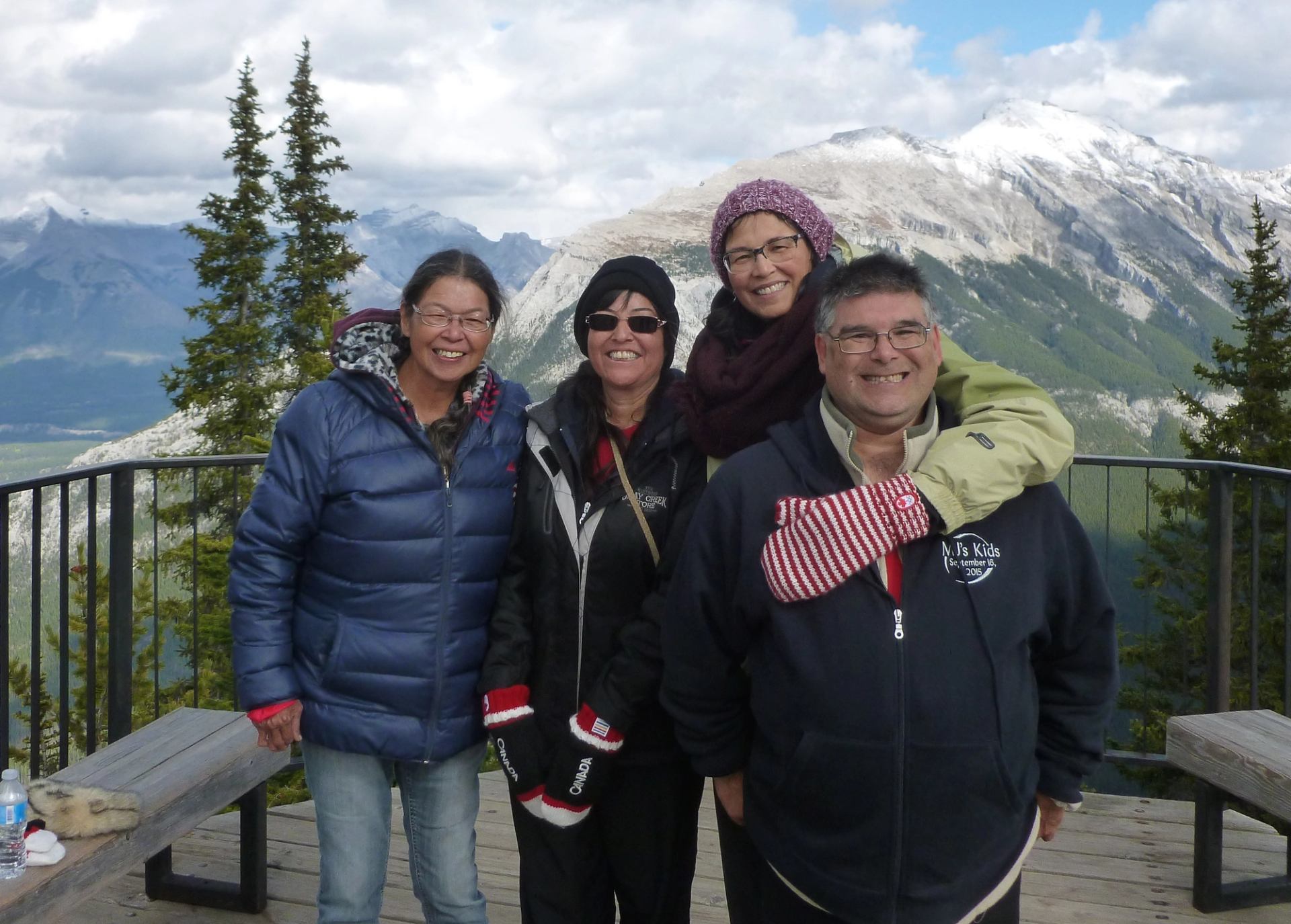 Four people in winter clothing stand on a mountain lookout with snowy peaks behind.