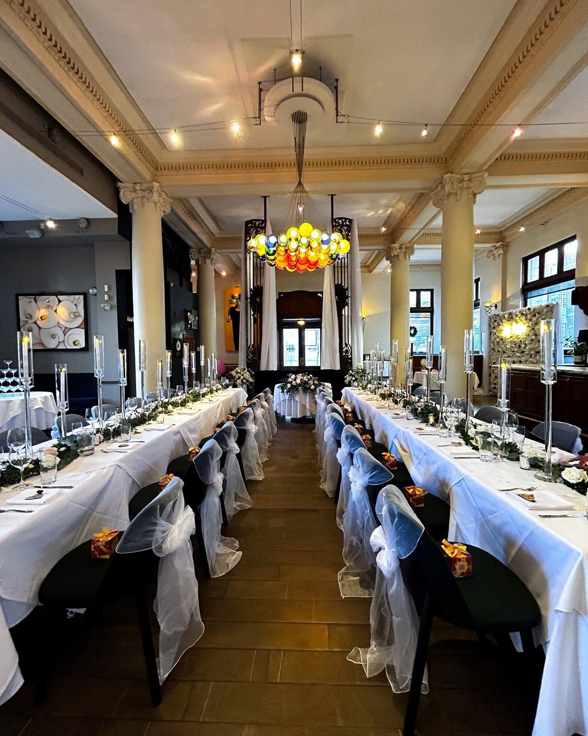 Formal banquet hall with long decorated tables, sheer chair covers and colourful chandelier.