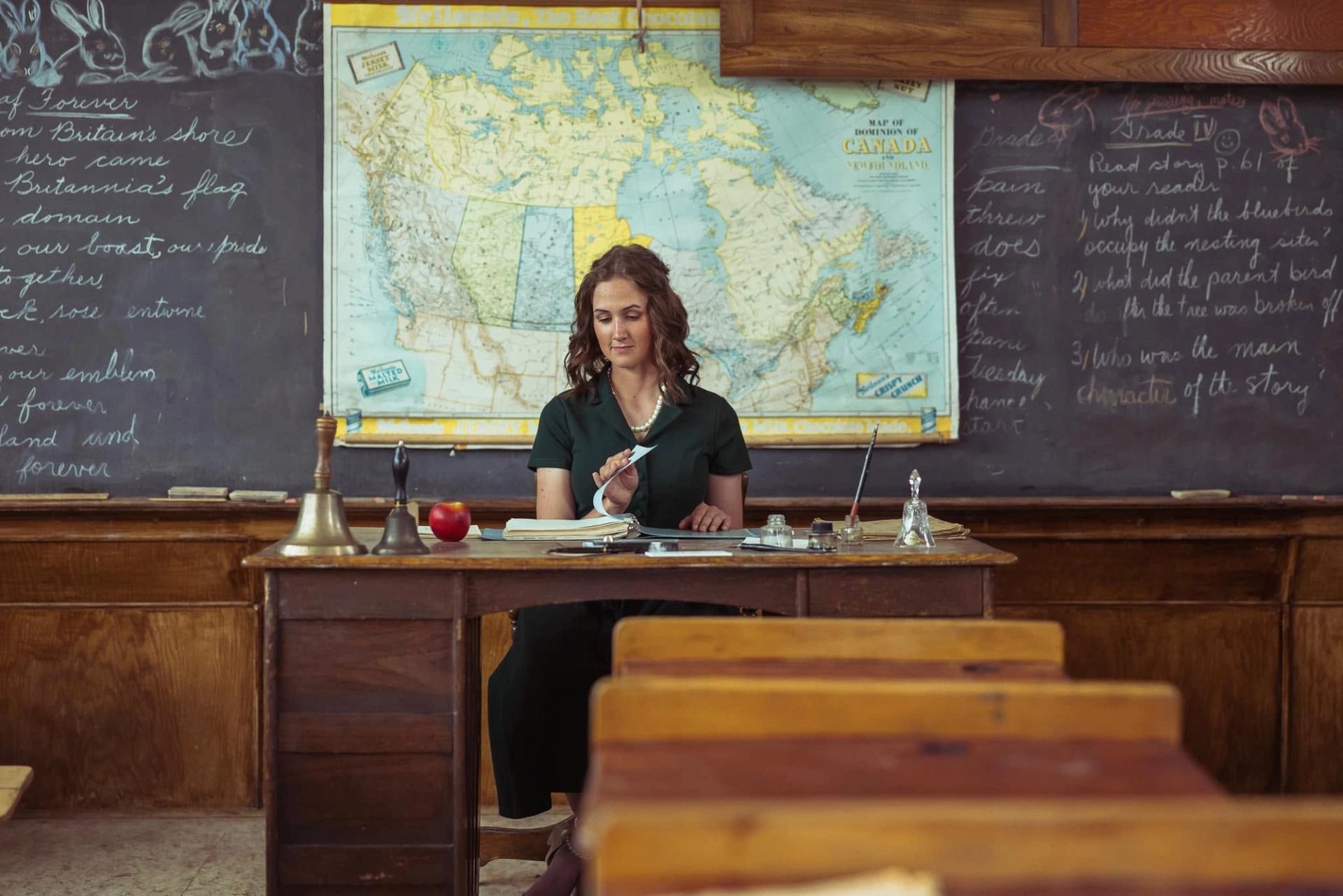 Teacher turns pages at a desk before a Canada map and chalkboards.