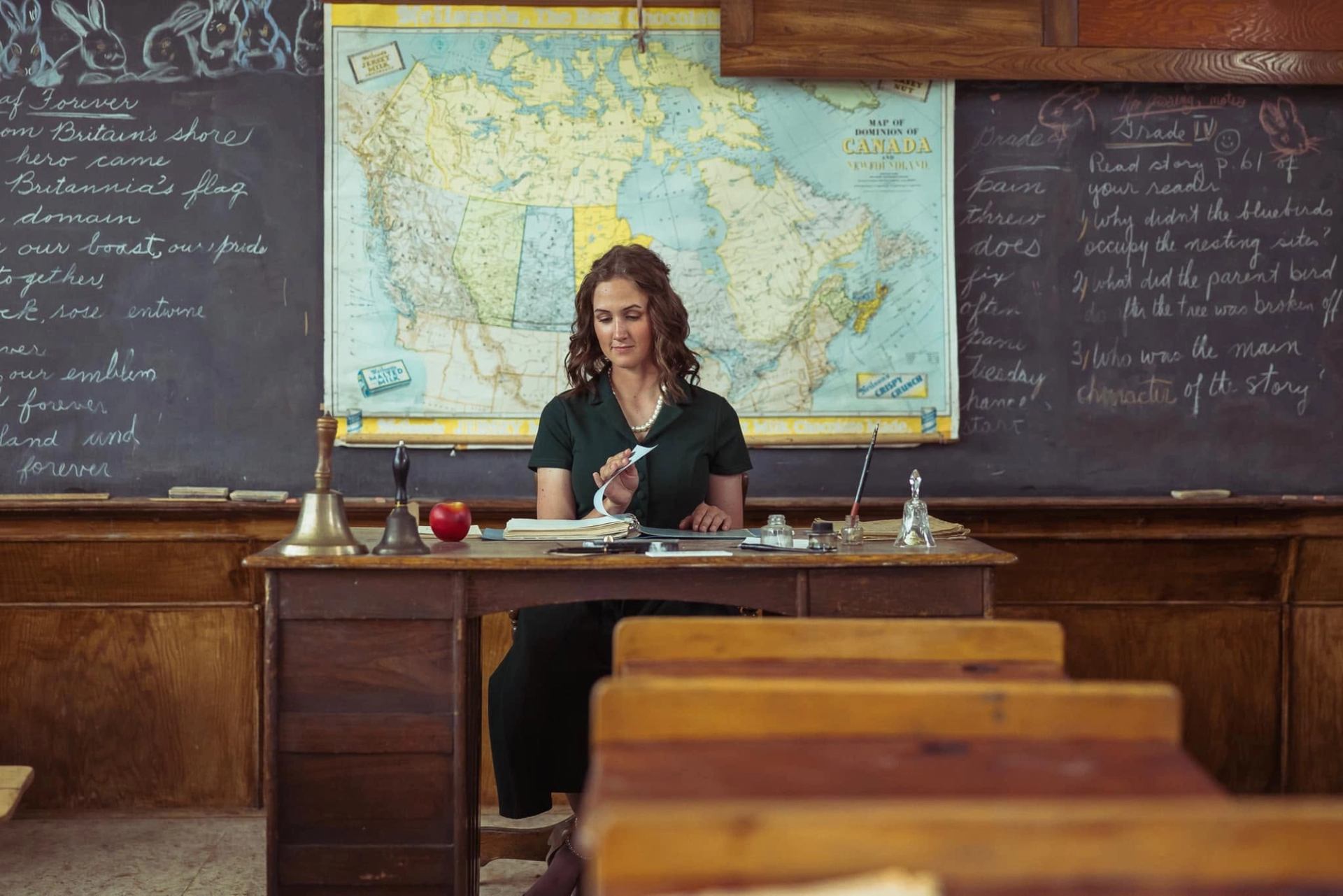 Teacher turns pages at a desk before a Canada map and chalkboards.