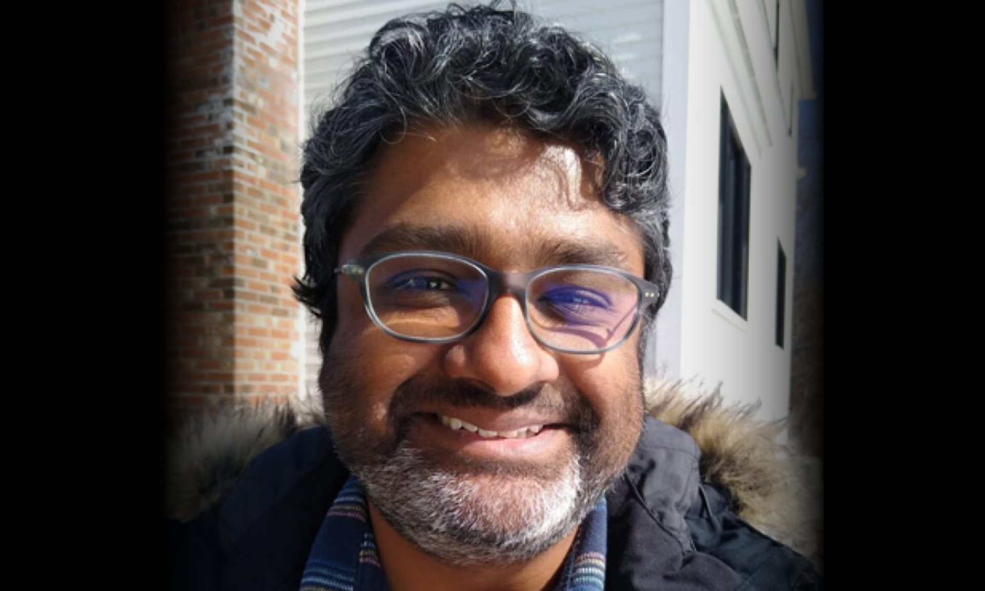 A smiling man with curly dark hair, a beard, and glasses wears a dark jacket with a fur-lined collar.