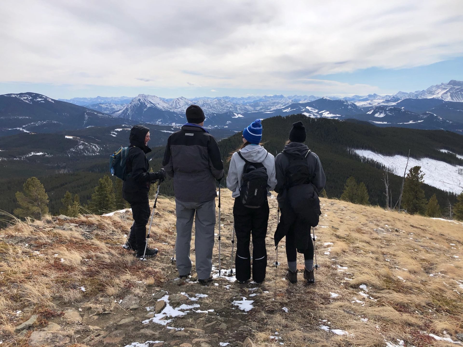 Four hikers stand on a ridge overlooking snowy mountains and valleys.