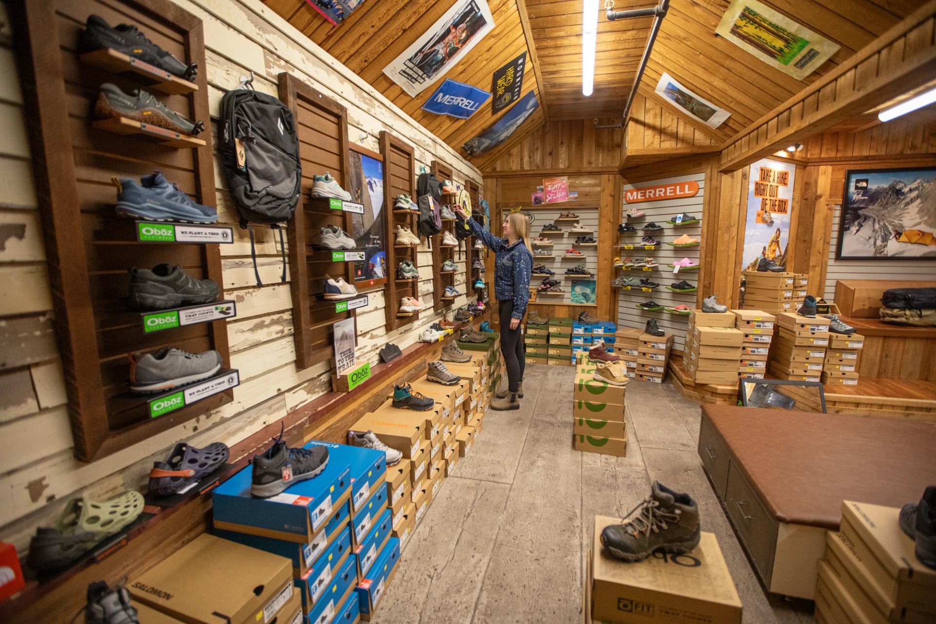 Outdoor shoe store interior with person browsing hiking footwear displayed on walls and boxes.