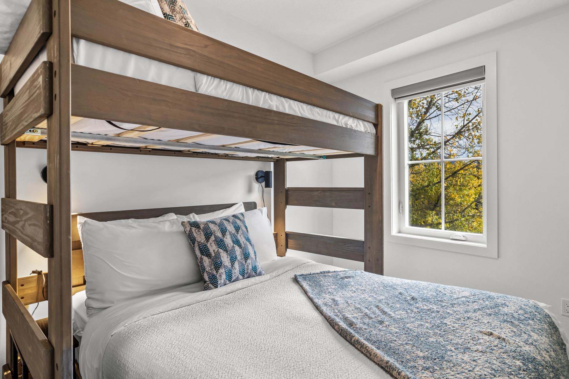 Wooden bunk bed with white bedding beside window showing leafy trees outside.